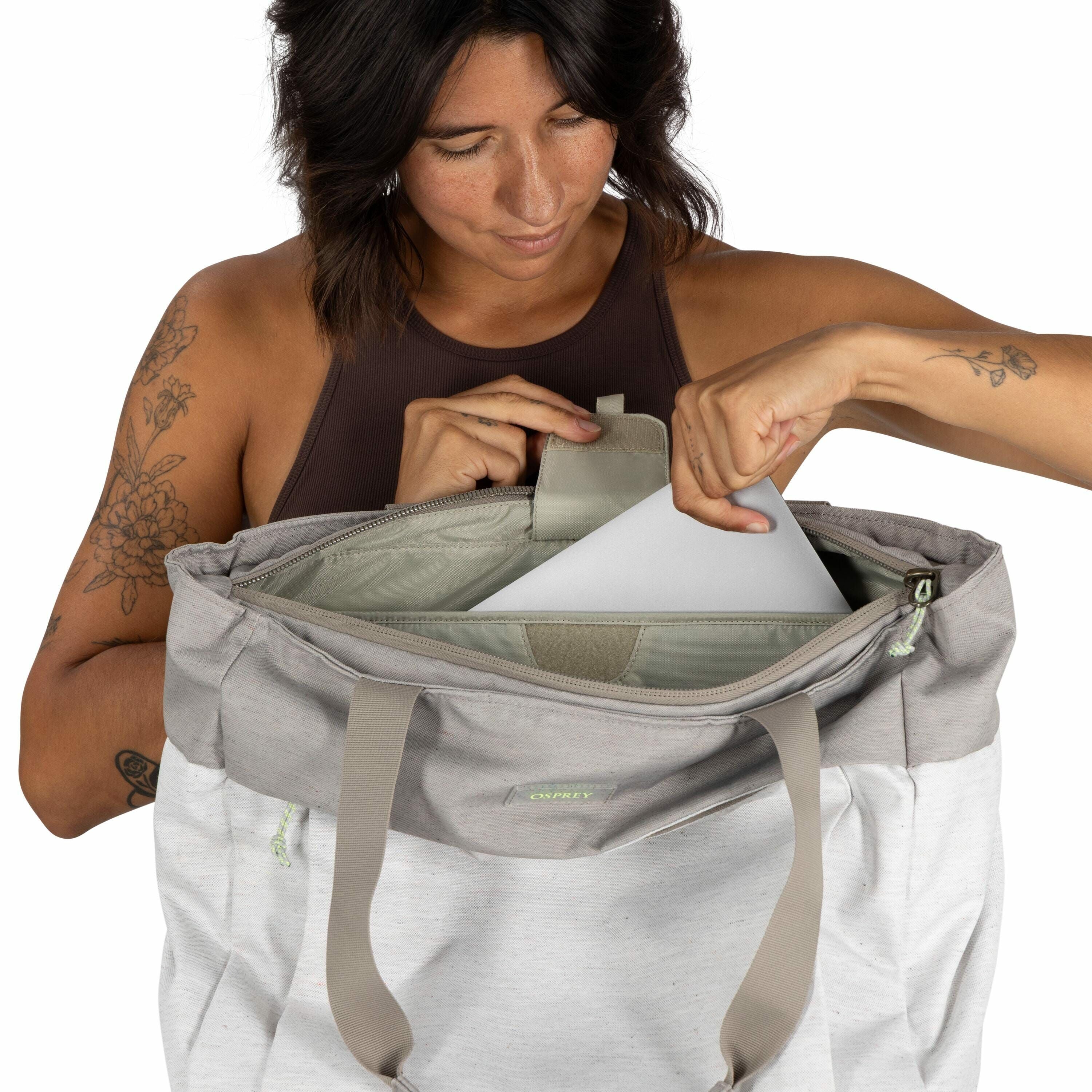 The image features a person wearing a sleeveless, dark brown top, revealing tattoos on both arms. The left arm has tattoos of flowers, while the right arm features a tattoo of a single rose with leaves. The individual is holding an Osprey branded bag, whi