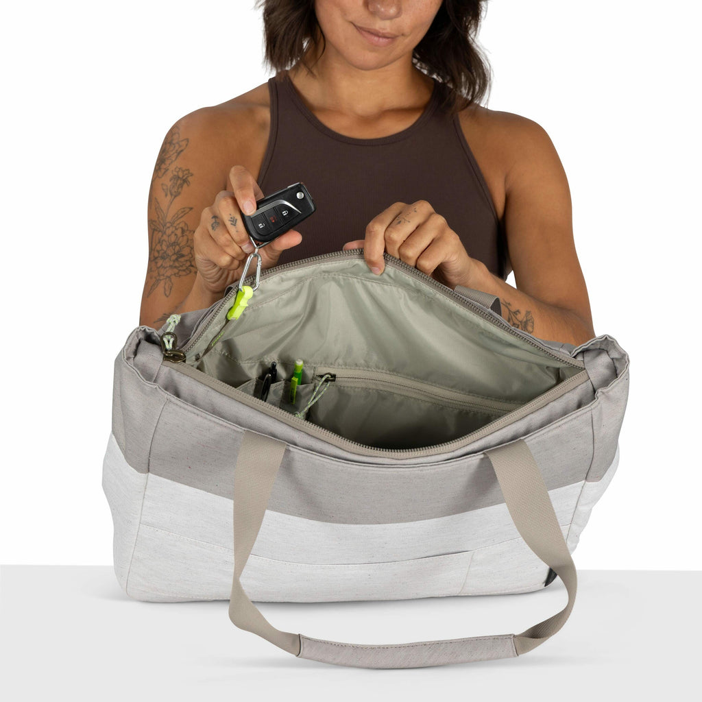 The image shows a woman with short brown hair, wearing a sleeveless brown top, holding a gray and white canvas bag. She is looking down at the bag, which is open, displaying a neatly organized interior with compartments holding items like pens. Her tattoo