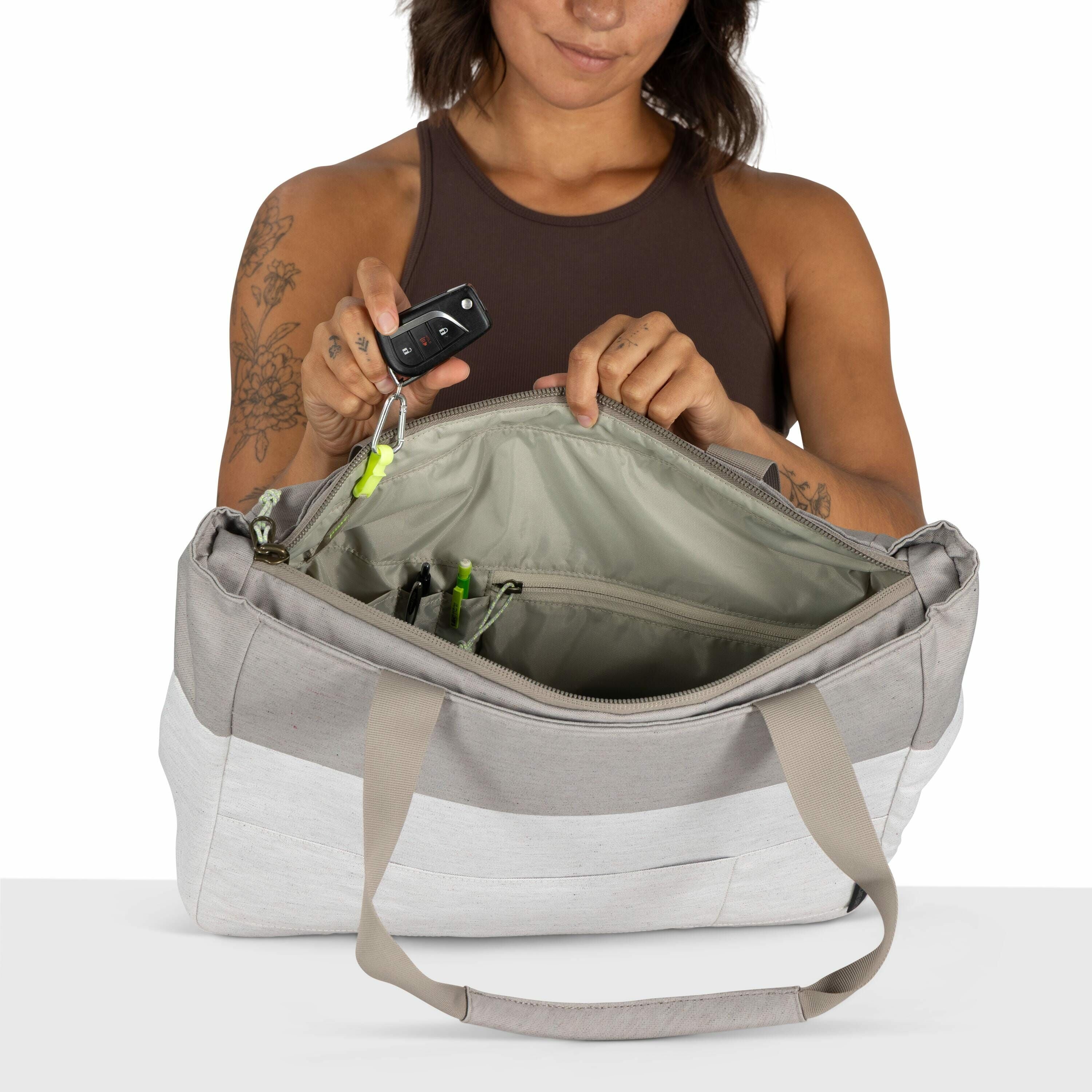 The image shows a woman with short brown hair, wearing a sleeveless brown top, holding a gray and white canvas bag. She is looking down at the bag, which is open, displaying a neatly organized interior with compartments holding items like pens. Her tattoo