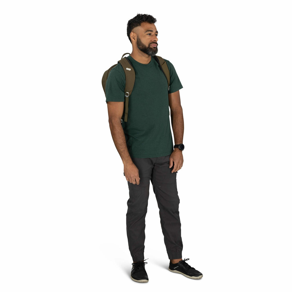 The image shows a man standing in a casual pose, wearing a dark green T-shirt and dark gray cargo pants. He has a small brown backpack on his shoulders, suggesting he might be prepared for a casual outing or a day hike. On his wrist, he is wearing a black