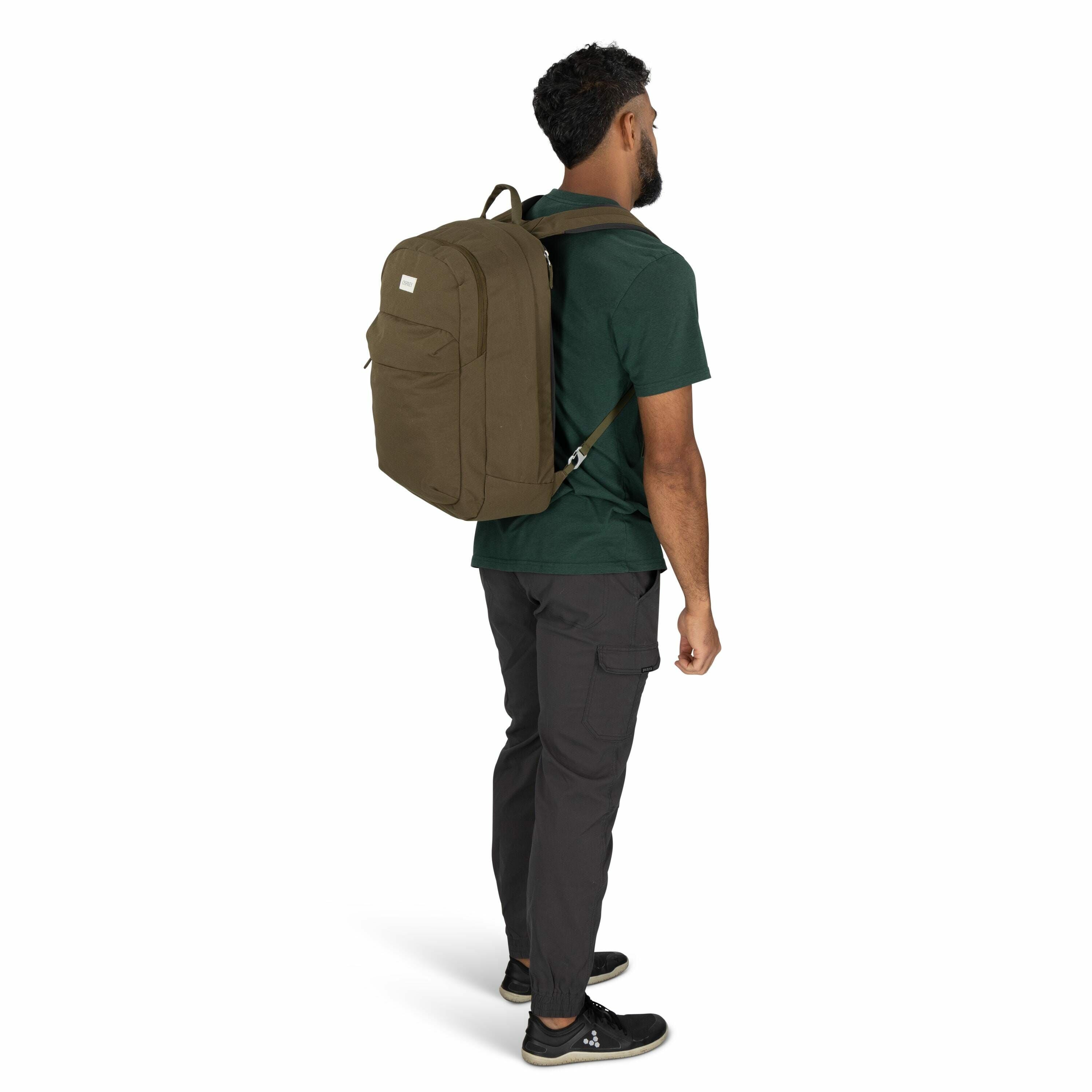 The image shows a man standing with his back to the camera, wearing a large olive green backpack. He is dressed in a dark green t-shirt and dark gray pants that have cargo pockets, and he is wearing black shoes. The backpack appears to be roomy and is sec