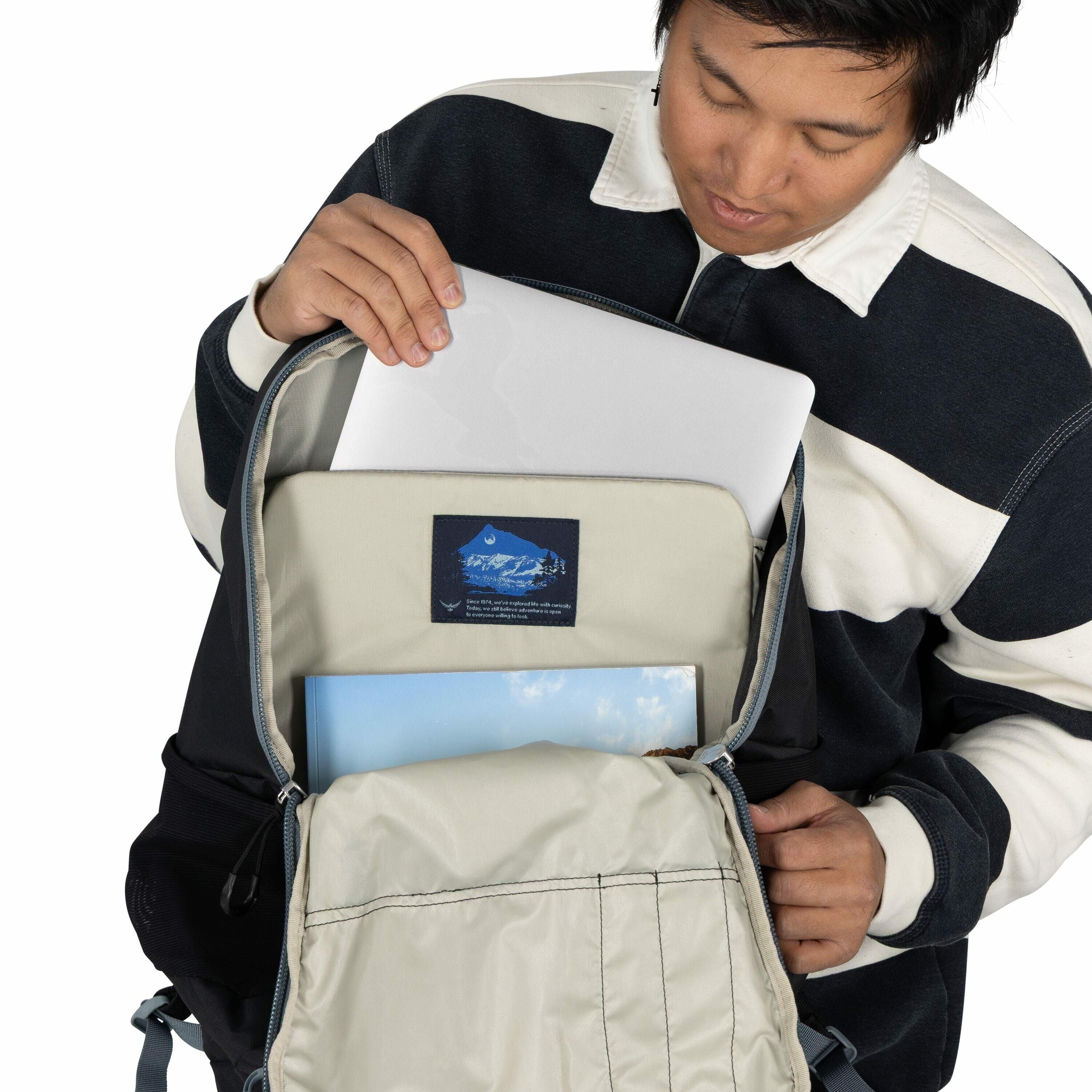 The image shows a person wearing a dark blue and white striped sweater holding a backpack that is partially opened. Inside the backpack, there is a laptop that the person is placing into a dedicated compartment. Below the laptop, there is a book with a sk