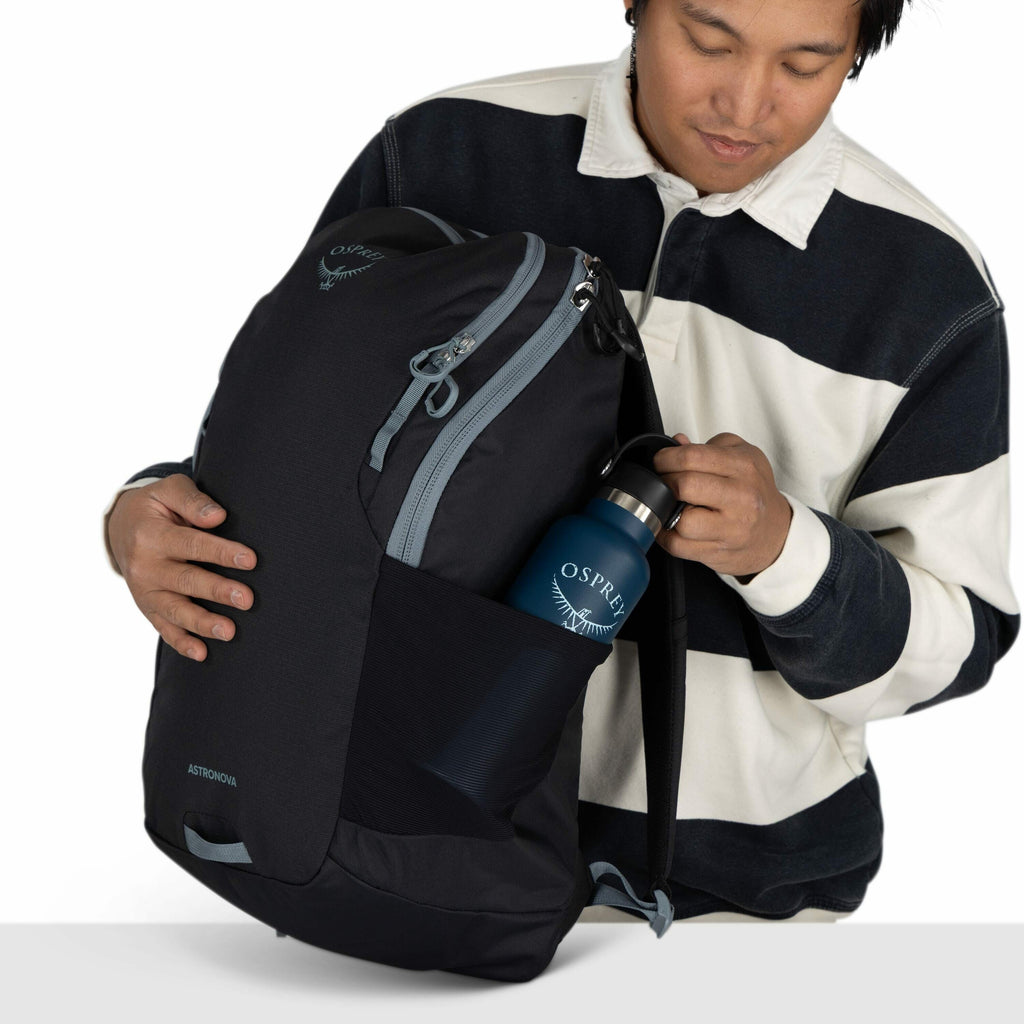 The image shows a person holding a black backpack branded with the logo "Osprey" near the top. The backpack features a small zippered compartment at the front and has a larger main compartment with gray zippers. The person is also holding a blue water bot
