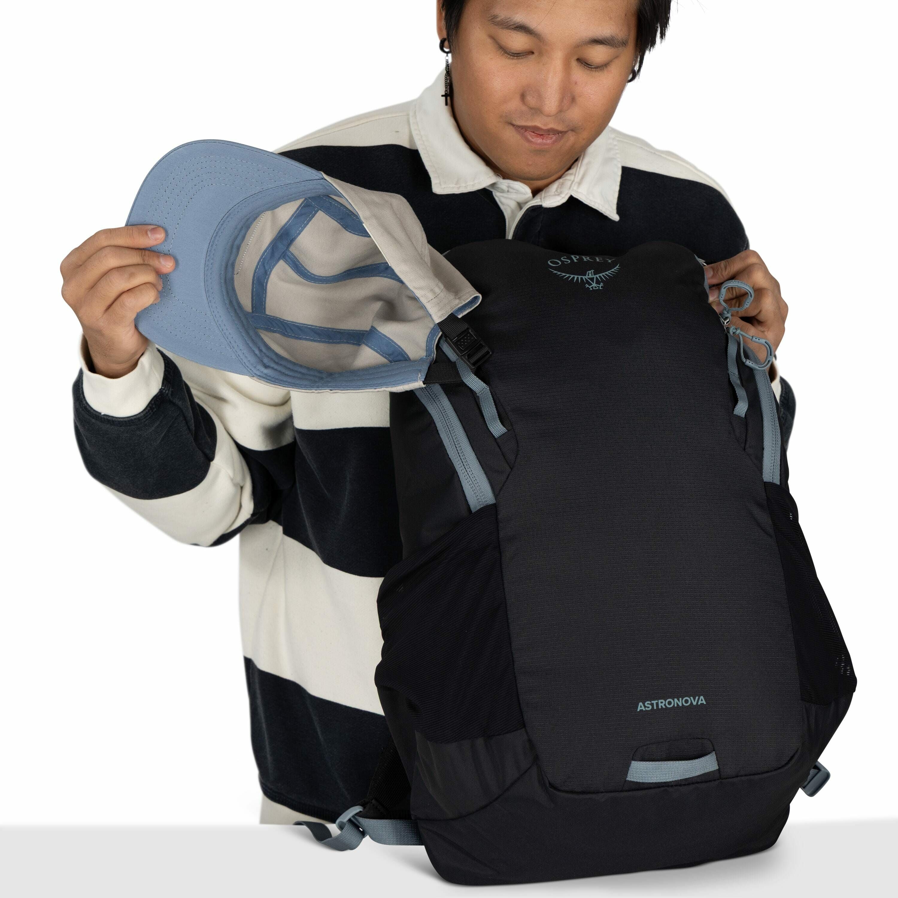 The image shows a person wearing a black and white striped sweater, holding a light blue baseball cap in one hand and a black backpack in the other. The backpack is branded with the logo "Osprey" and the model name "Astronova" printed in white. The backpa