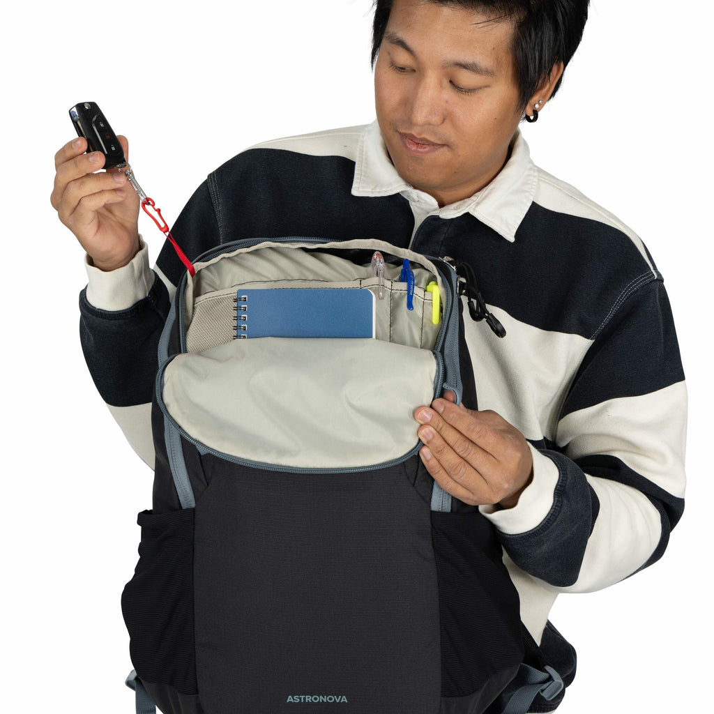 The image shows a person holding open a backpack from the brand Astronova. The backpack's main compartment is visible, containing various items such as a blue notebook, pens, a highlighter, and other office supplies. The person is also holding a car key,