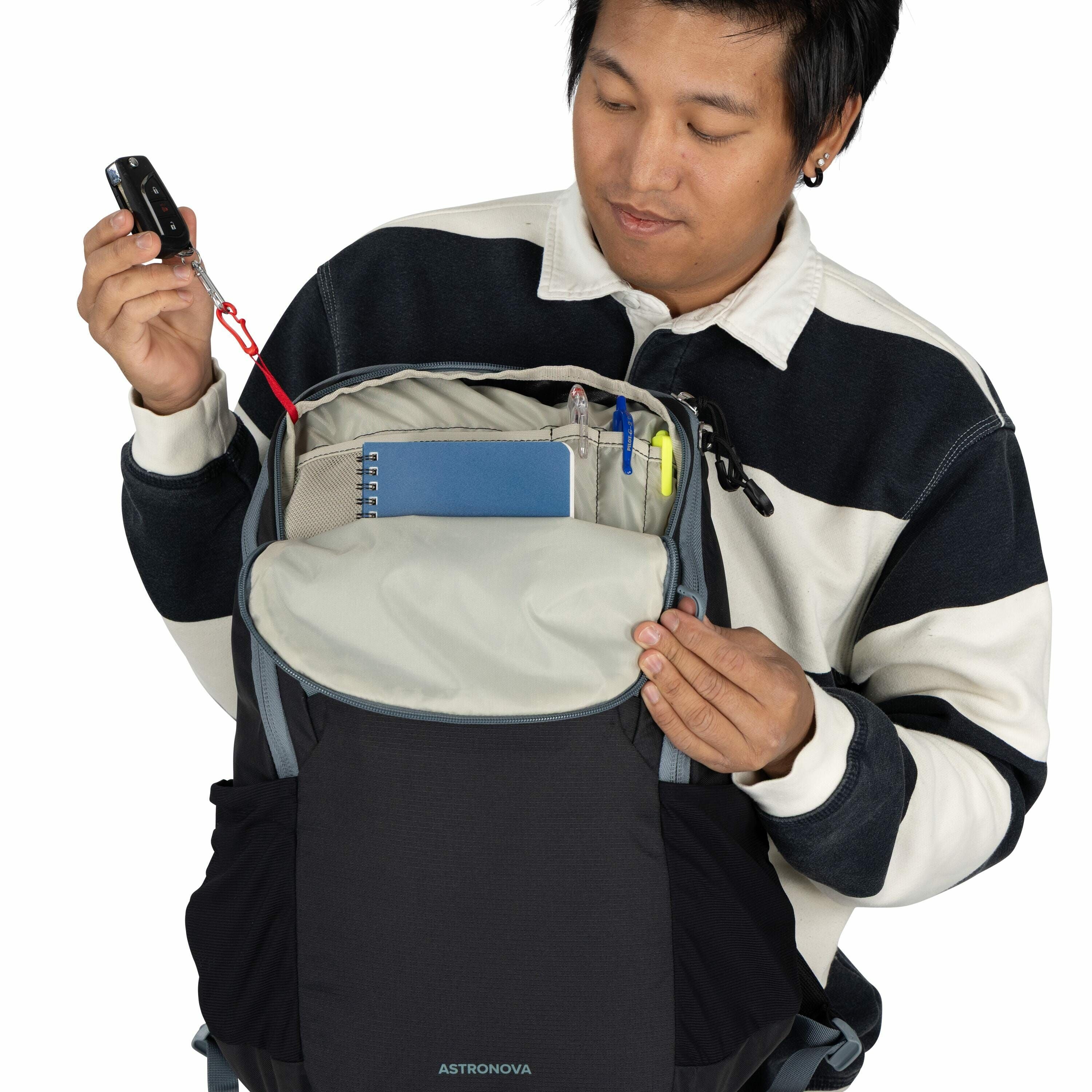 The image shows a person holding open a backpack from the brand Astronova. The backpack's main compartment is visible, containing various items such as a blue notebook, pens, a highlighter, and other office supplies. The person is also holding a car key,