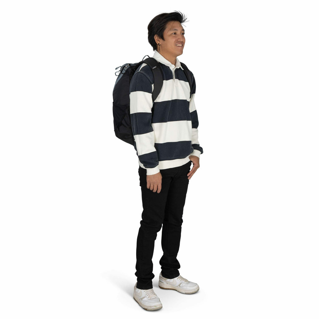 The image depicts a person standing and facing slightly to the side. They are wearing a casual outfit consisting of a long-sleeved rugby shirt with thick horizontal navy and white stripes, black pants, and white sneakers. On their back is a large black ba