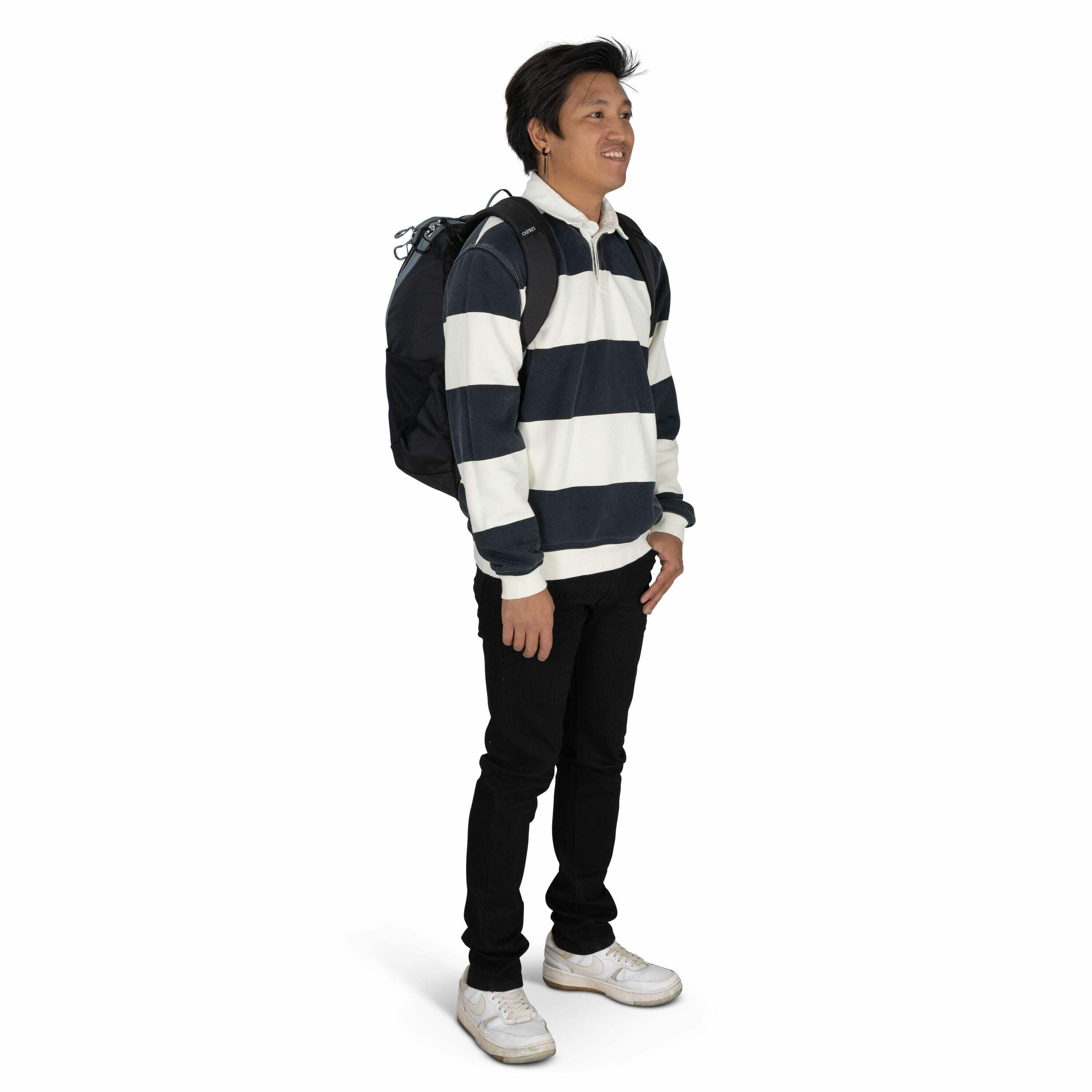 The image depicts a person standing and facing slightly to the side. They are wearing a casual outfit consisting of a long-sleeved rugby shirt with thick horizontal navy and white stripes, black pants, and white sneakers. On their back is a large black ba
