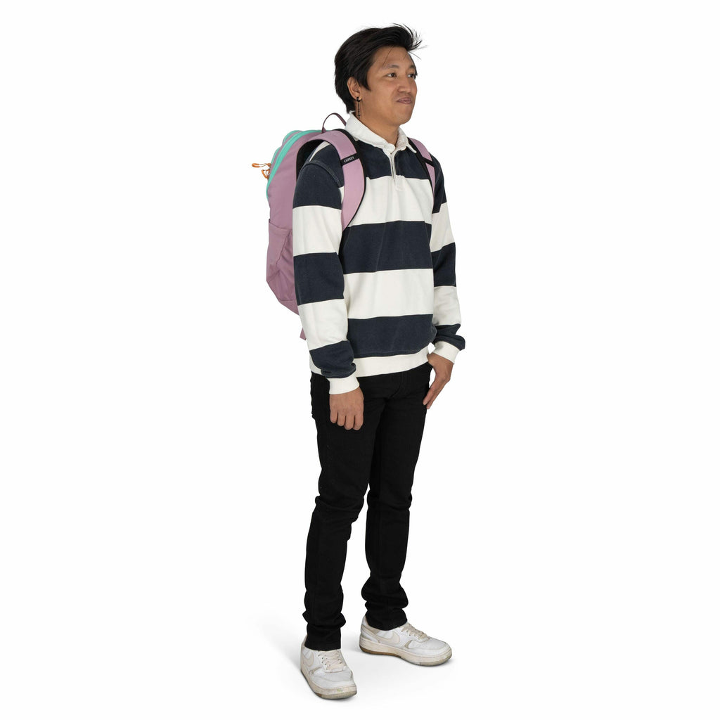The image shows a person standing with a relaxed posture, wearing a black and white striped long-sleeve polo shirt, black pants, and white sneakers. They are carrying a light purple backpack with pastel-colored accents on their back. Their hair is short a