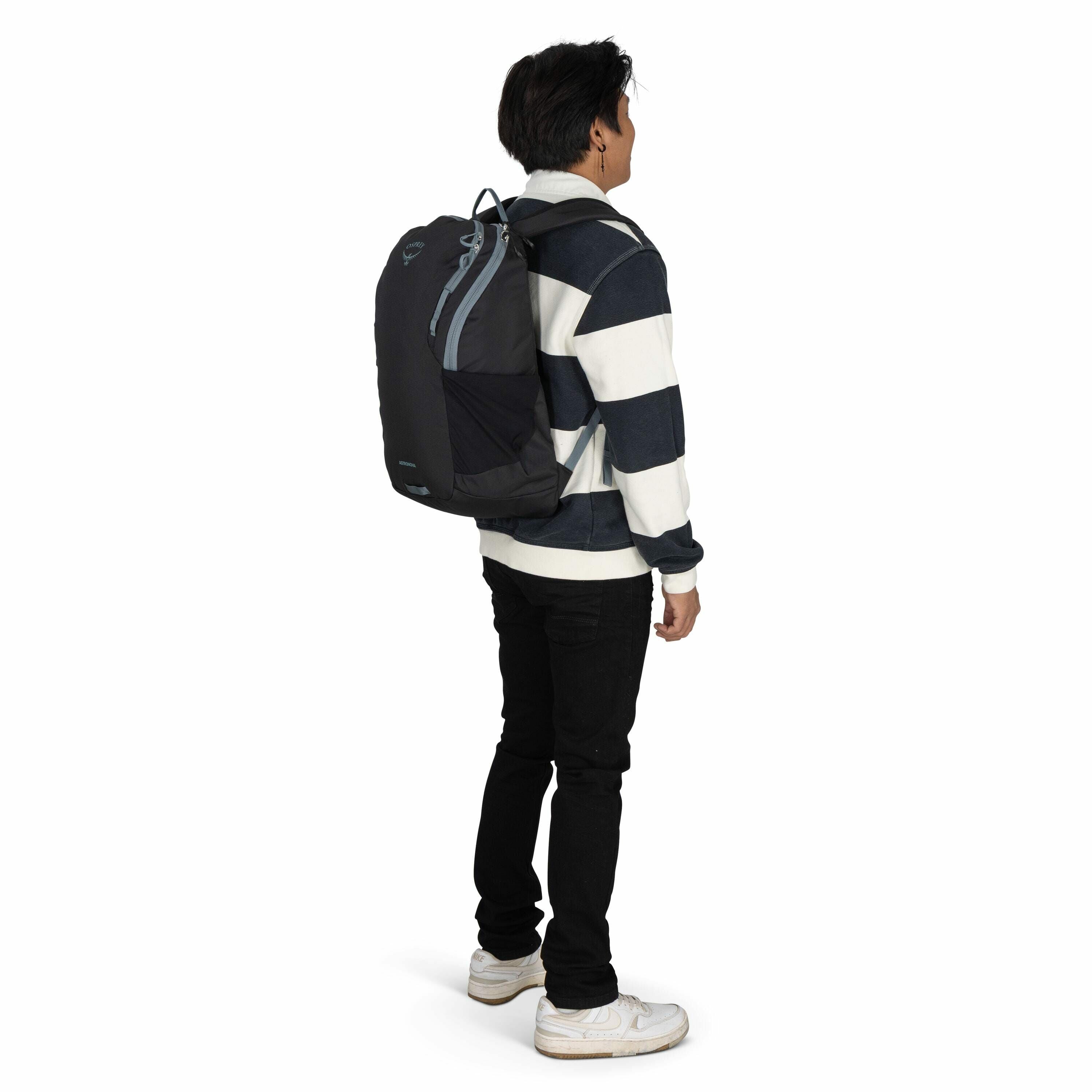 The image features a young man facing slightly to the side, wearing a black and white striped rugby shirt and a pair of black pants. He is carrying a large black backpack with light blue accents on his back, which appears to be an Osprey brand, as indicat