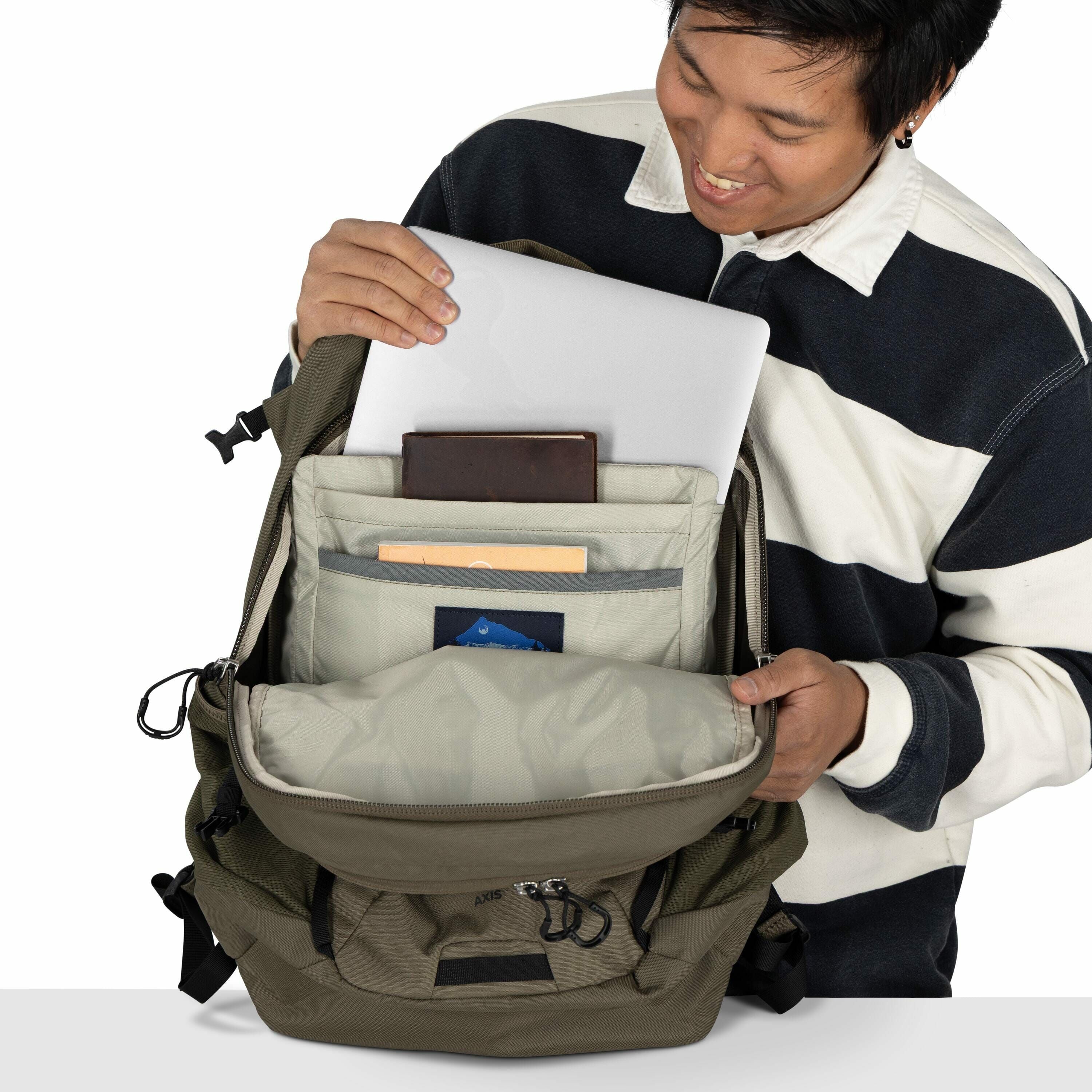 In the image, a person is holding open a khaki-colored backpack, displaying its organized contents. Inside the backpack, there is a padded compartment that holds a silver laptop, indicating the backpack is designed for electronics protection. Additionally
