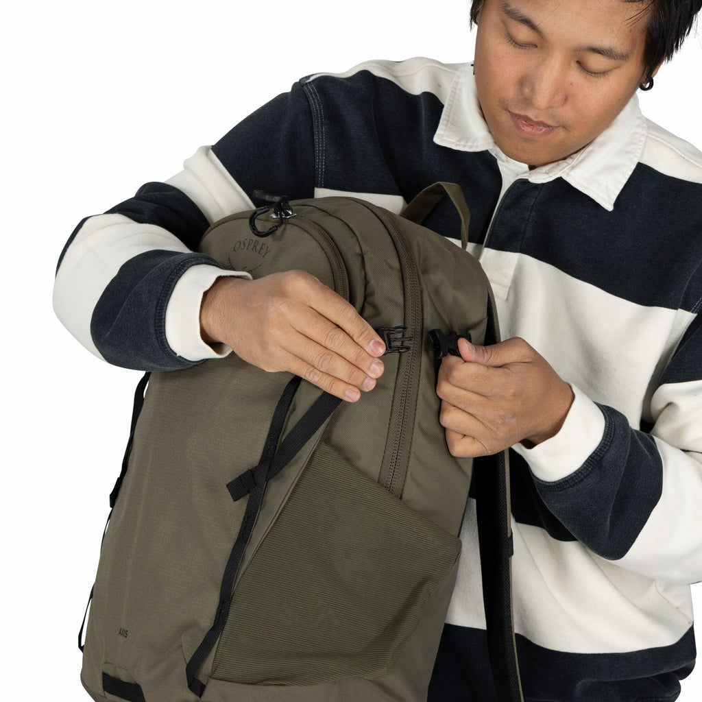 The image shows a person wearing a dark blue and white striped sweater with a white collar. The individual is holding an olive green Osprey backpack, adjusting or fastening one of its straps or buckles. The backpack appears to be a practical and durable m
