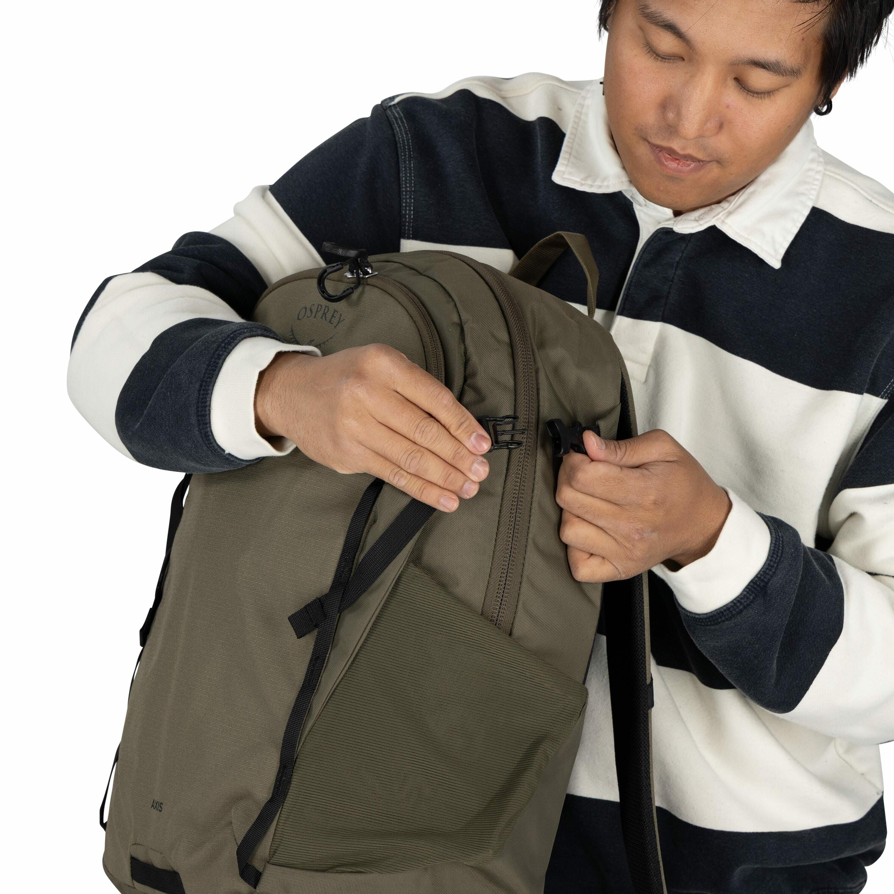 The image shows a person wearing a dark blue and white striped sweater with a white collar. The individual is holding an olive green Osprey backpack, adjusting or fastening one of its straps or buckles. The backpack appears to be a practical and durable m