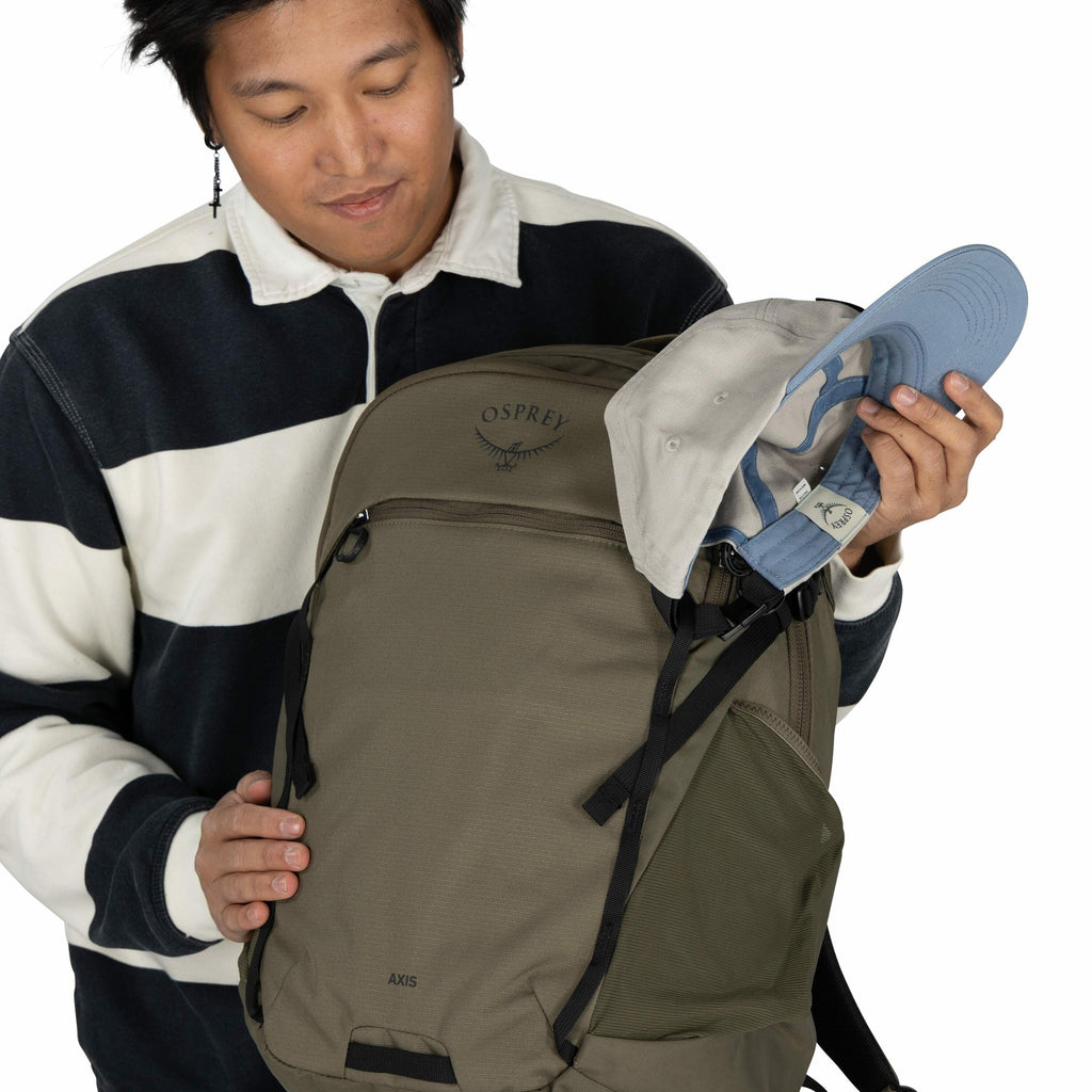 The image shows a person wearing a black and white striped long-sleeve shirt. They are holding an Osprey backpack, which is olive green with black straps, and has the "OSPREY" logo with an axis design. The person is also holding a light gray and blue base