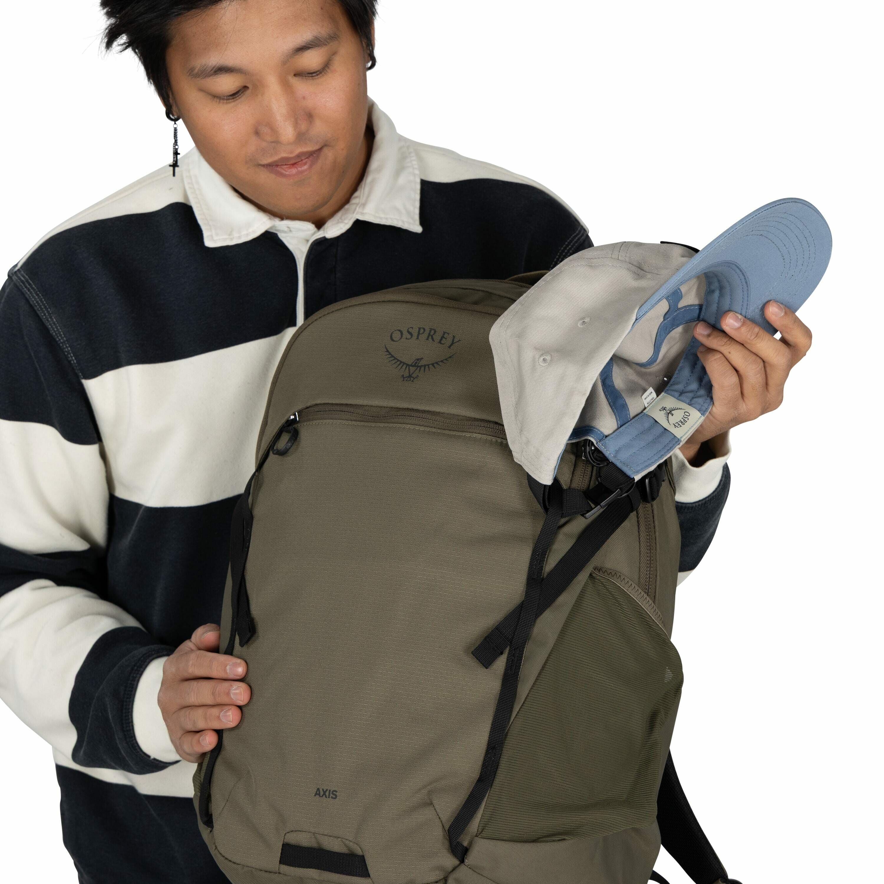 The image shows a person wearing a black and white striped long-sleeve shirt. They are holding an Osprey backpack, which is olive green with black straps, and has the "OSPREY" logo with an axis design. The person is also holding a light gray and blue base