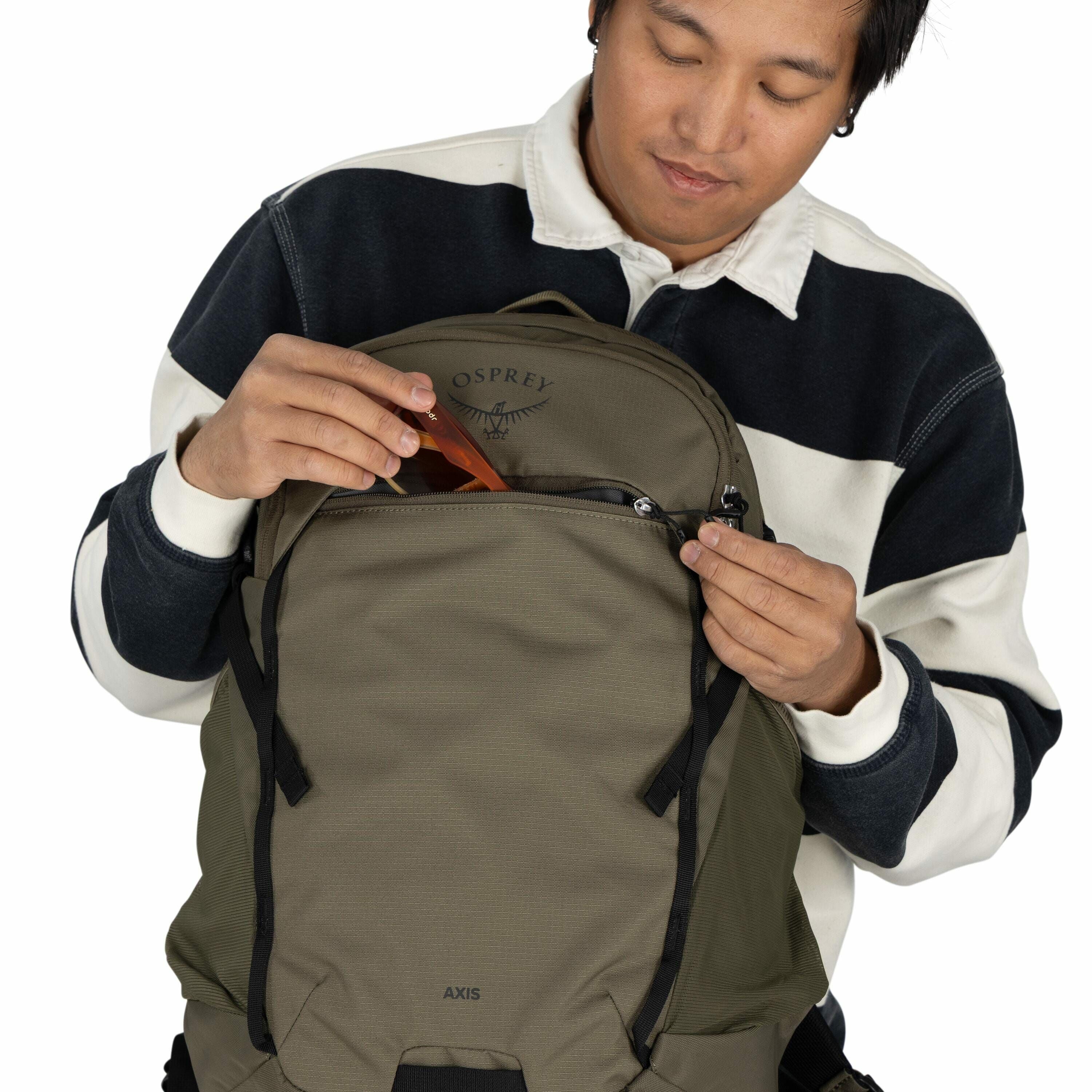 The image shows a person wearing a striped sweater in black, white, and grey tones. They are holding an olive-green Osprey Axis backpack, with a visible logo on the upper part of the bag. The person is in the process of placing or removing an orange item,