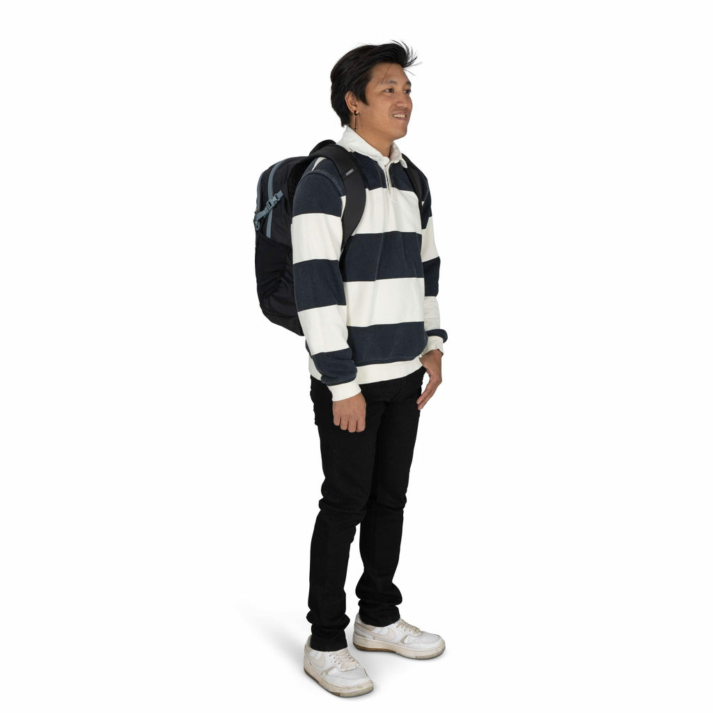 The image depicts a person standing and facing slightly to the left, carrying a dark-colored backpack on their back. The individual is wearing a long-sleeved rugby-style shirt with broad horizontal stripes in navy blue and white. They have paired this shi