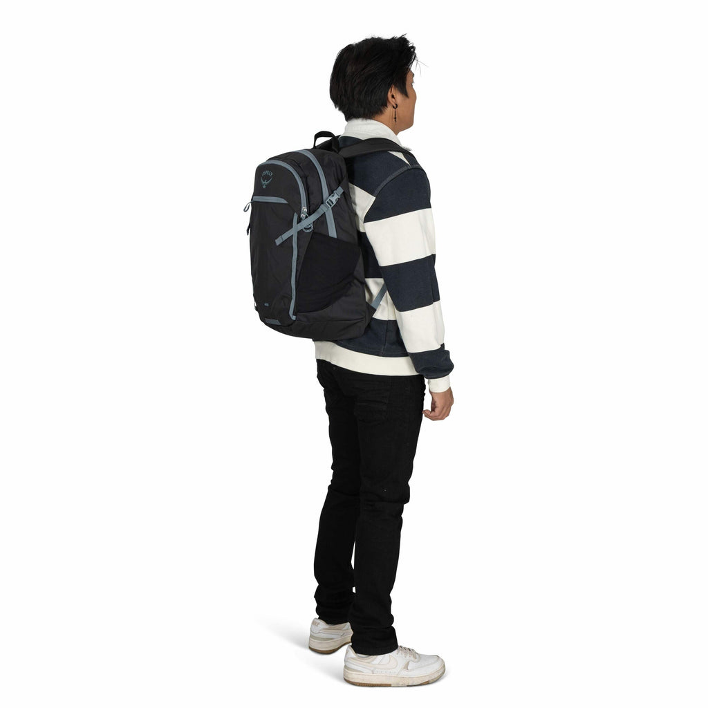 The image features a young man standing in a side profile view, wearing a black and white striped sweater, black pants, and white sneakers. He is carrying a dark-colored Osprey backpack with light blue accents on his back. The backpack appears to be well-