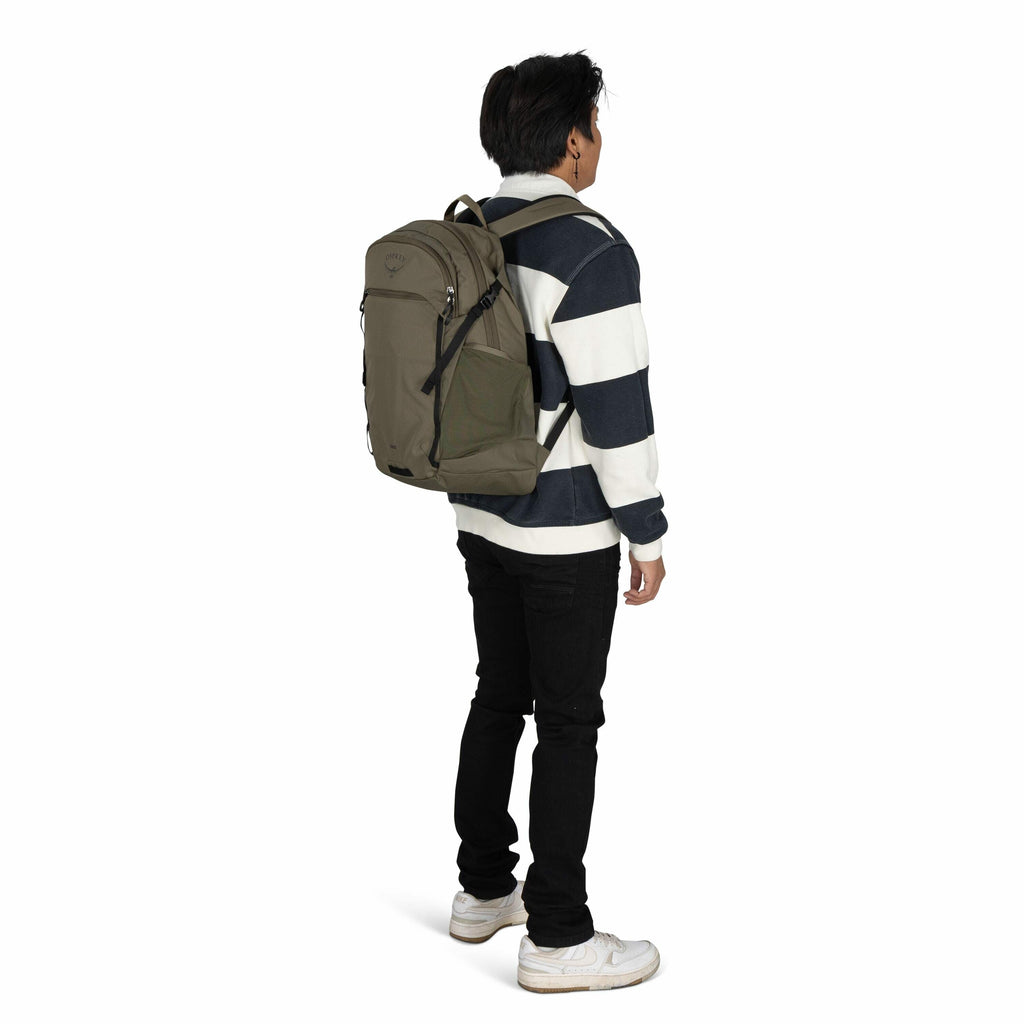 The image features a young person standing and facing away from the camera, wearing a large, olive-green backpack. The backpack has the "Osprey" logo visible on the upper section. The individual is dressed casually in a black and white striped sweater, bl