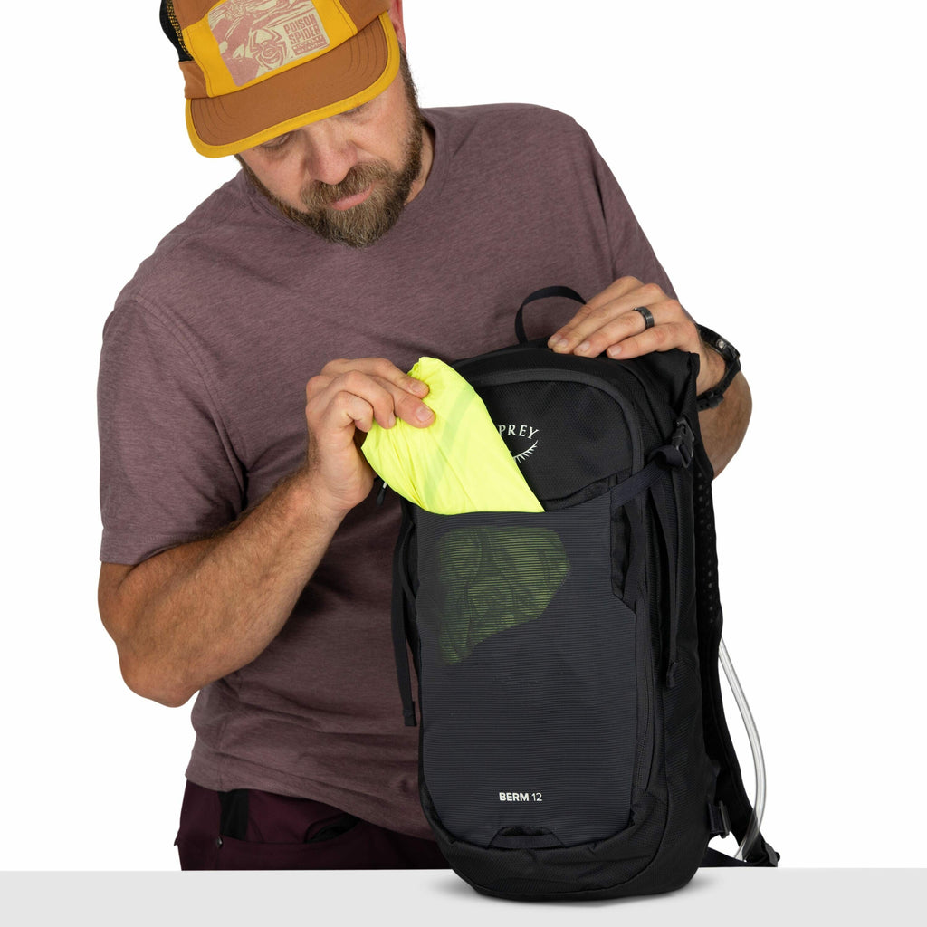 The image shows a man wearing a brown short-sleeved t-shirt and burgundy pants. He is holding a black Osprey backpack with the model name "Berm 12" printed on it. The man is inserting a folded neon yellow item, possibly a lightweight jacket or pouch, into