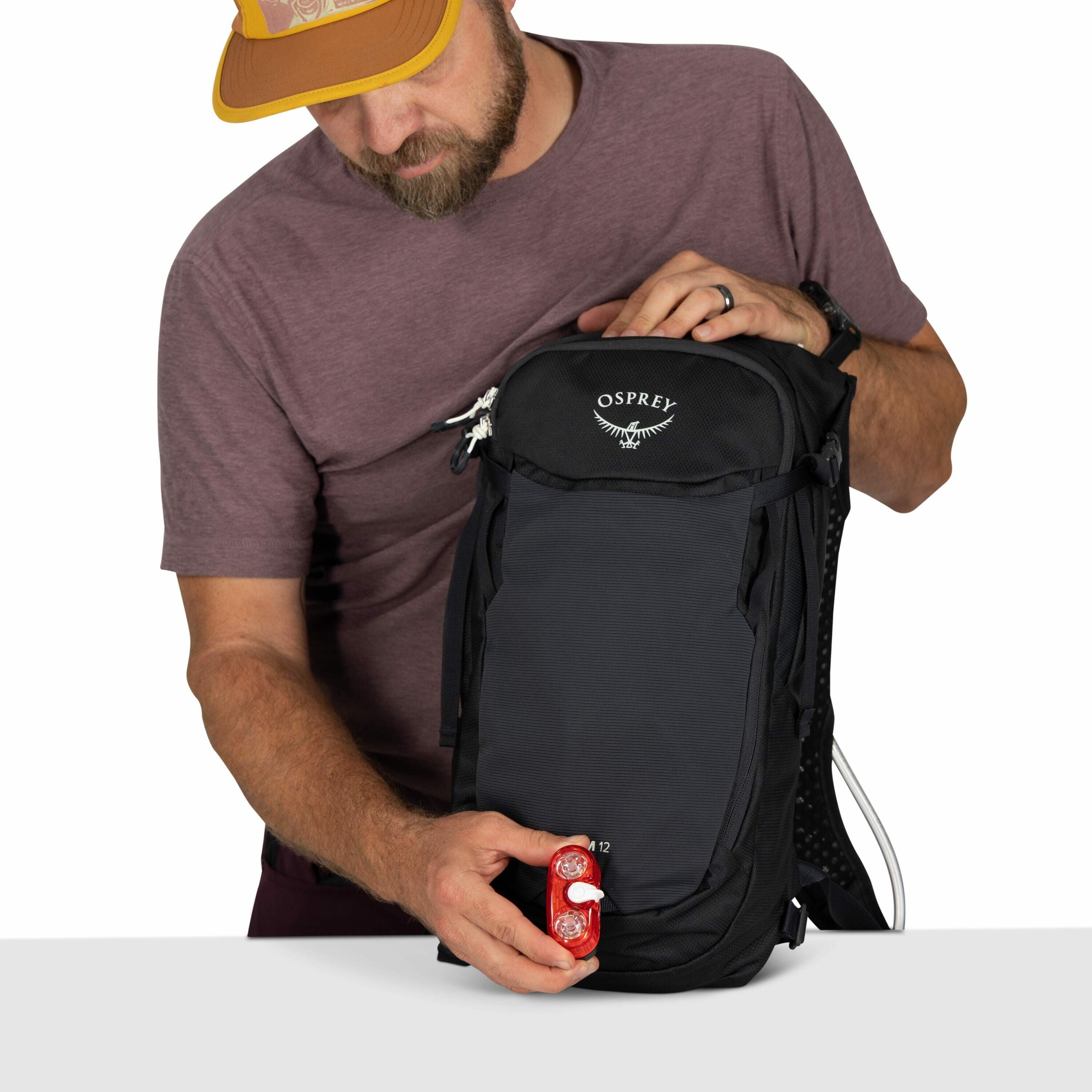 The image features a person dressed in a casual brown shirt, wearing a brown and yellow cap. They are holding a black Osprey backpack with both hands. In their left hand, they hold a small red bike light, which appears to be a safety accessory for cycling
