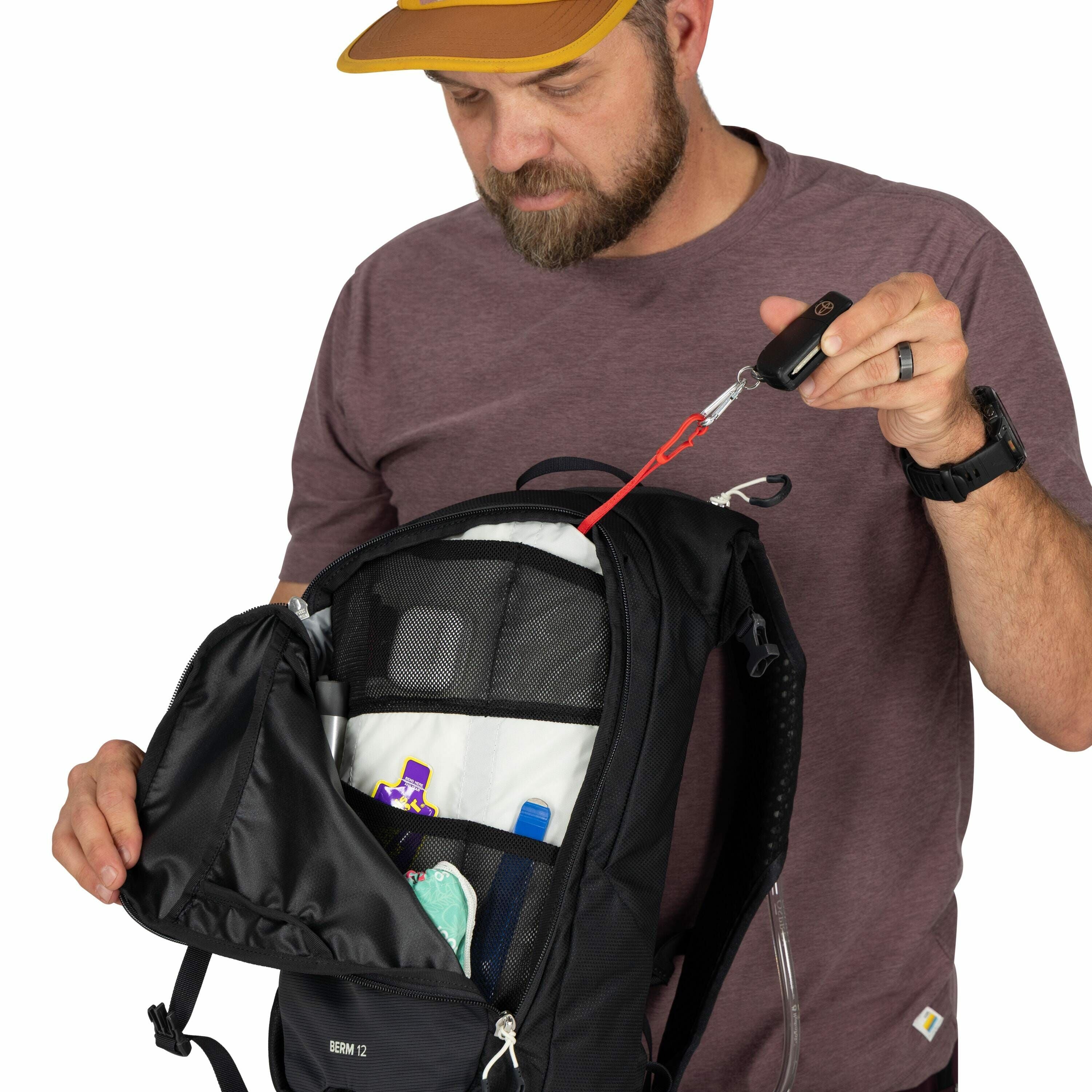 The image features a person wearing a casual dark-colored short-sleeve shirt, holding open a black backpack with several compartments visible inside. In the open backpack, there are various items including a purple and white packaged product, a teal objec