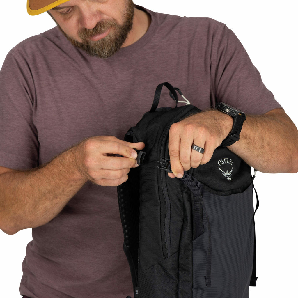 The image shows a person dressed in a purple t-shirt, wearing a watch on the left wrist and a ring on the right hand. The person is adjusting or attaching a black buckle on a black Osprey backpack. The backpack features visible branding, including the Osp