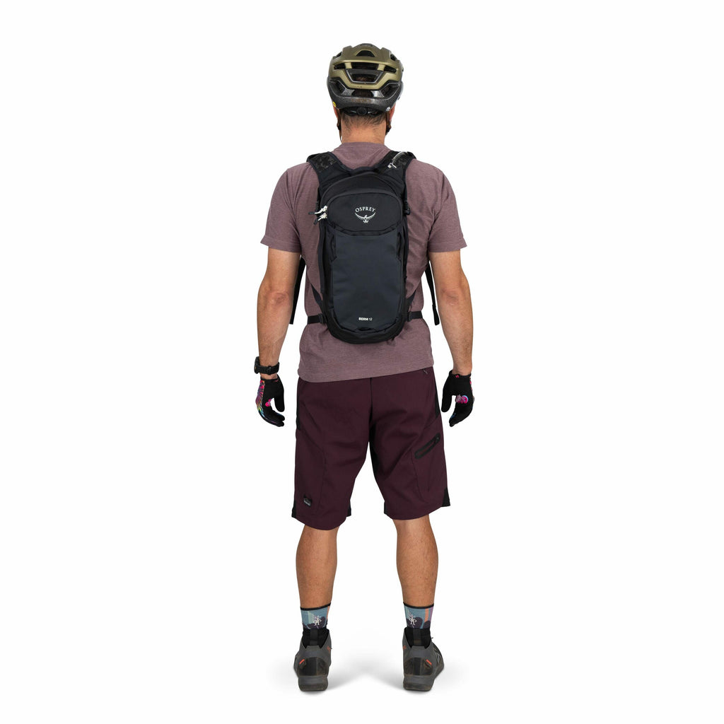 The image features a person standing with their back facing the camera, dressed in cycling attire. They are wearing a dark helmet and a black Osprey backpack with the Osprey logo clearly visible. The outfit includes a maroon short-sleeved shirt and maroon