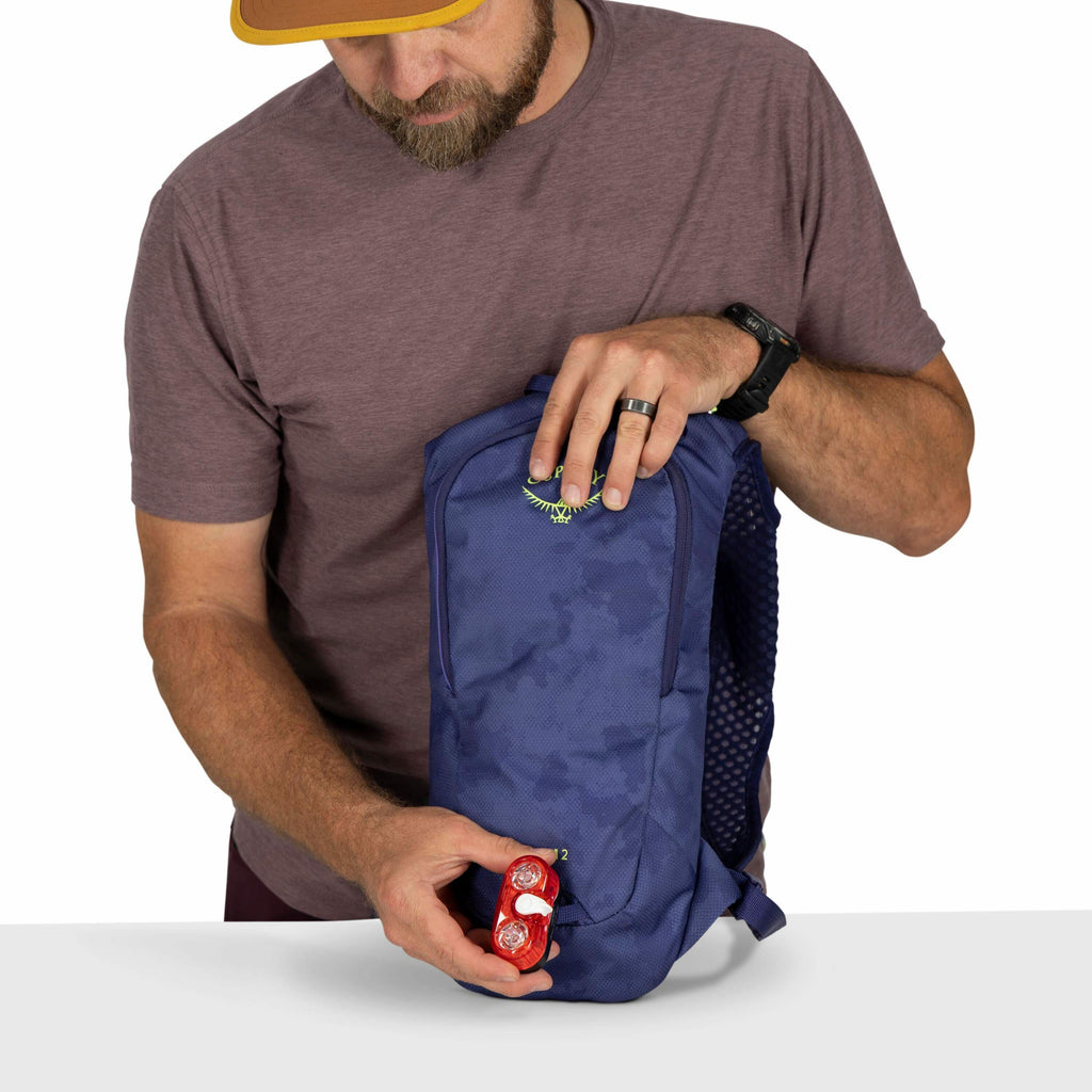 The image depicts a man holding a blue backpack from Osprey. He is wearing a maroon shirt and a watch on his left wrist, along with a black ring on his left hand. In his right hand, he holds a red object that appears to be a bike light or reflector. The b