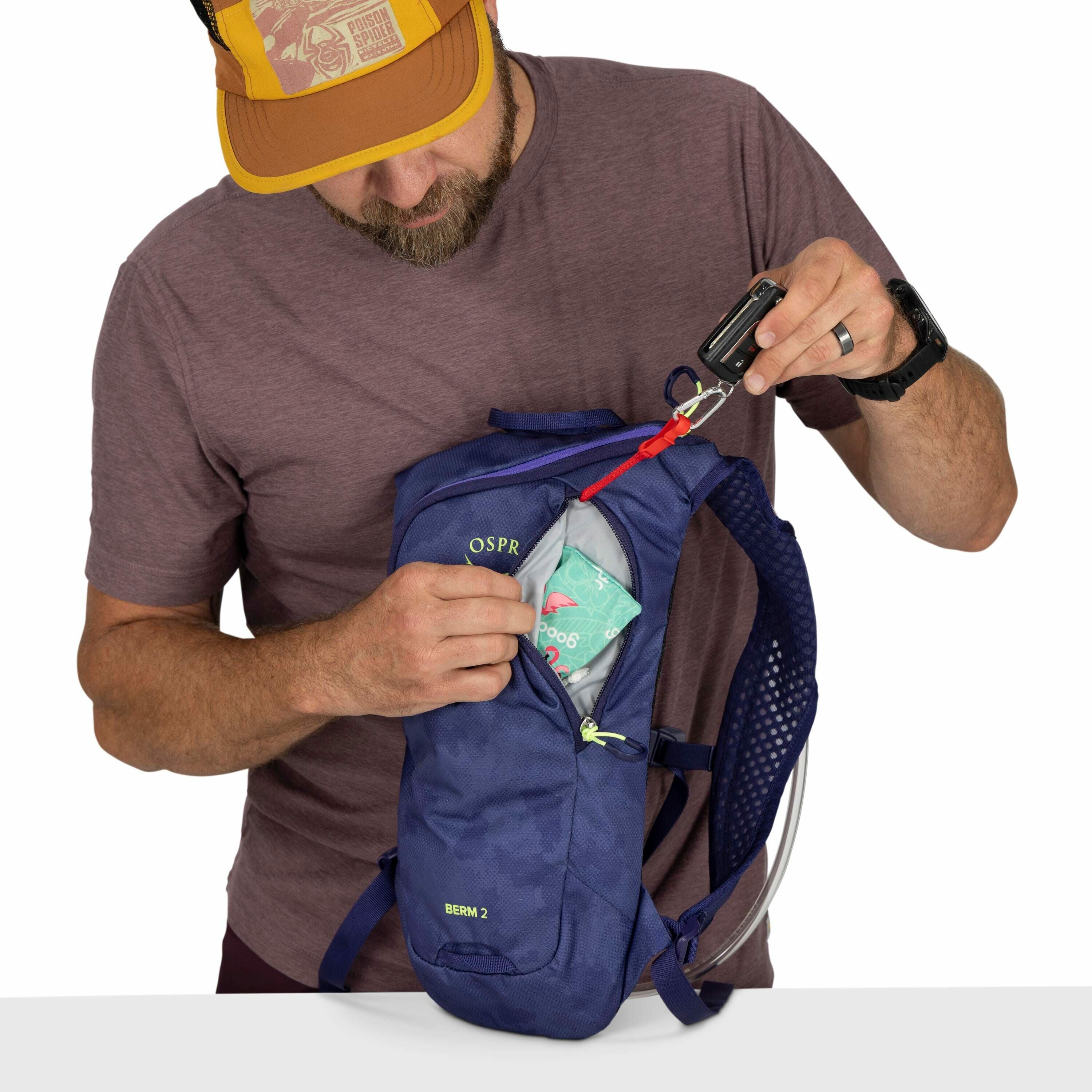 In the image, a person is holding a navy blue backpack labeled "OSPR" and "BERM 2" while wearing a maroon t-shirt and a mustard-yellow cap. The backpack is open, revealing a colorful package inside with pink and teal graphics and the number "206". The per