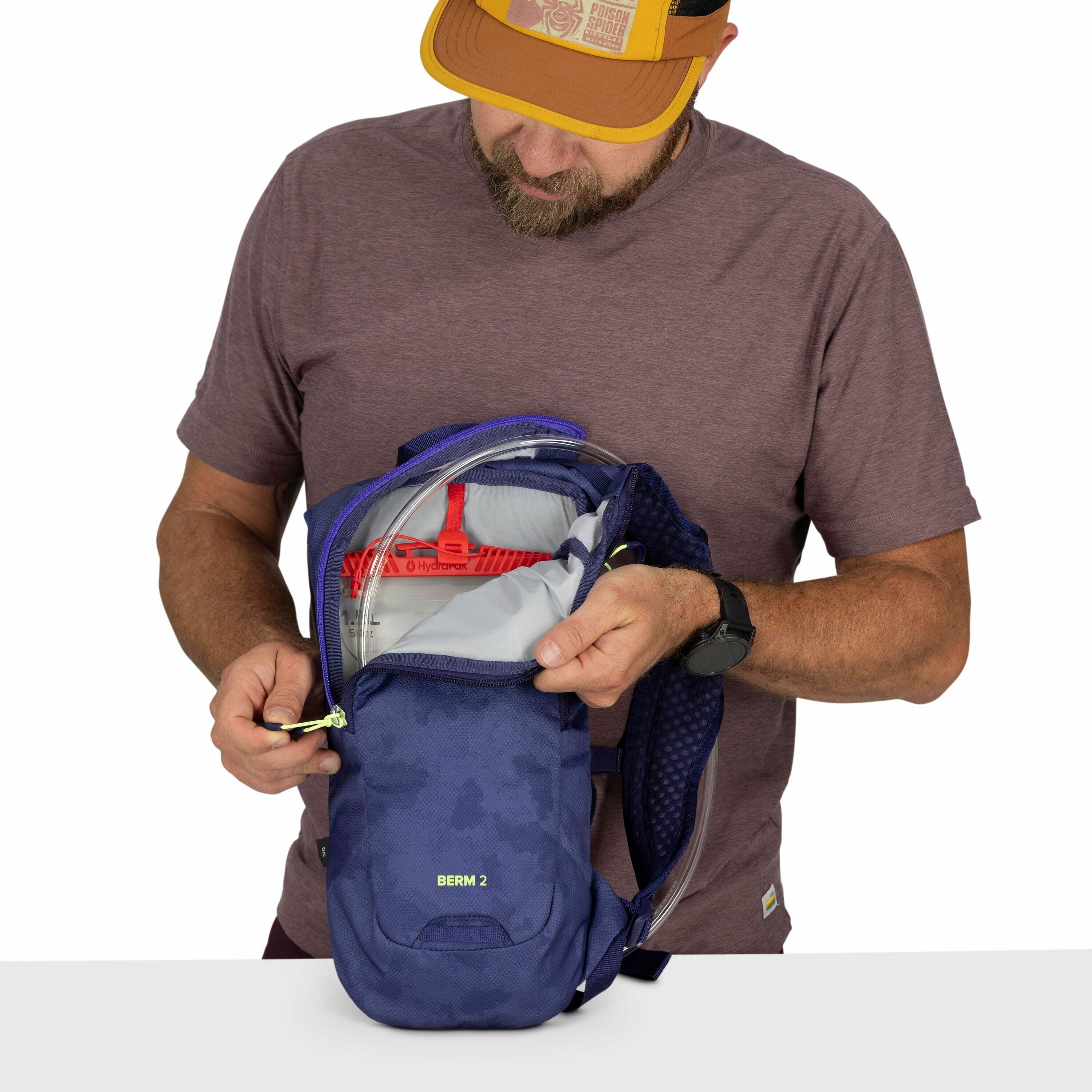 The image shows a person wearing a brown t-shirt and a yellow cap, holding a blue backpack. The backpack is open, revealing its contents, which include a red Hydrapak hydration reservoir system. The name "BERM 2" is visible on the front of the backpack, p