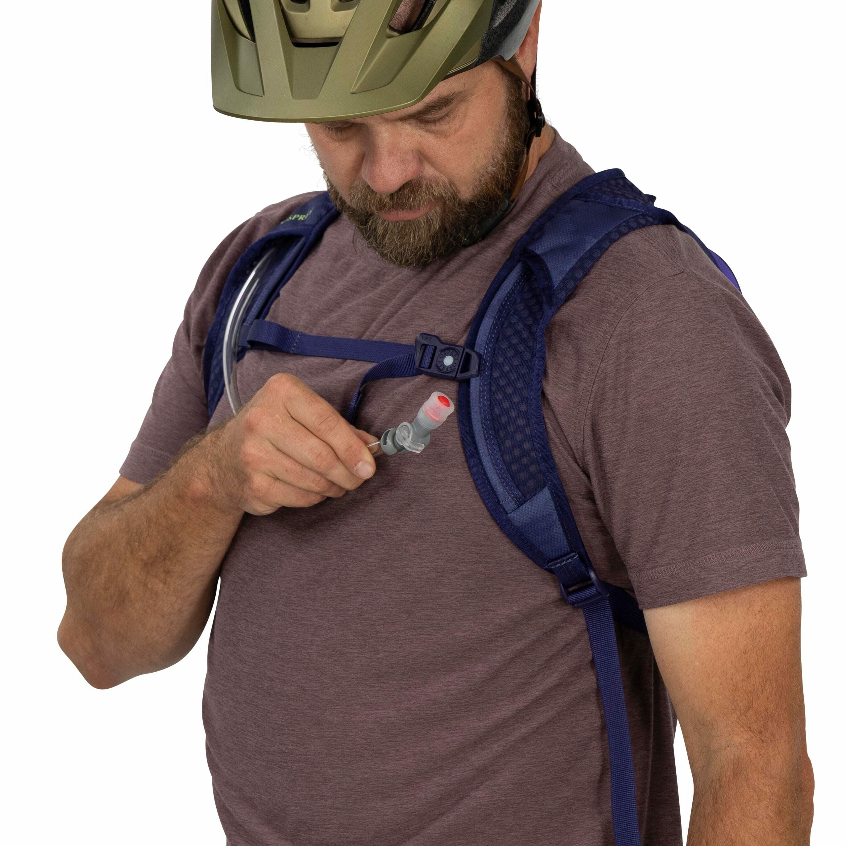The image depicts a person wearing a purple short-sleeve shirt and a blue hydration backpack. The backpack has adjustable straps and features a visible tube for drinking water. The individual is holding a bite valve connected to the tube, which is used to