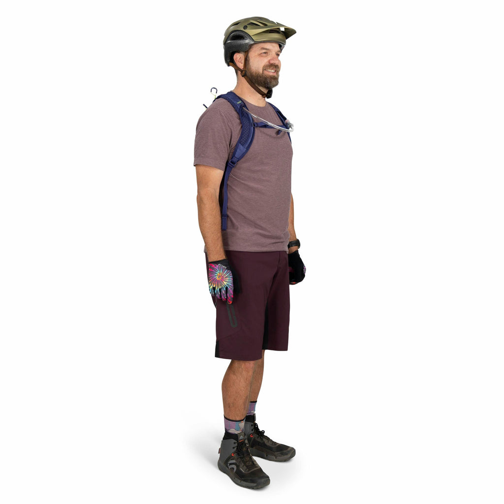 The image depicts a person standing in profile view dressed for biking or outdoor activities. They are wearing a gray helmet and a light purple t-shirt paired with burgundy shorts. On their hands, they have colorful gloves with a circular pattern that res