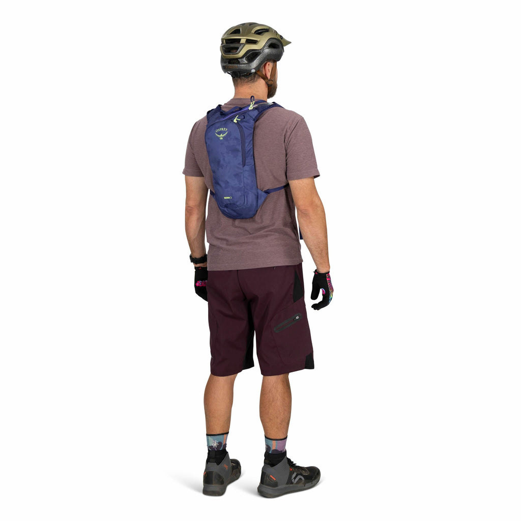 The image shows a person standing and facing away from the camera, wearing biking gear. They have a blue Osprey backpack on their back, a purple short-sleeve shirt, and dark shorts with a zippered pocket on the left side. The person is also wearing a helm