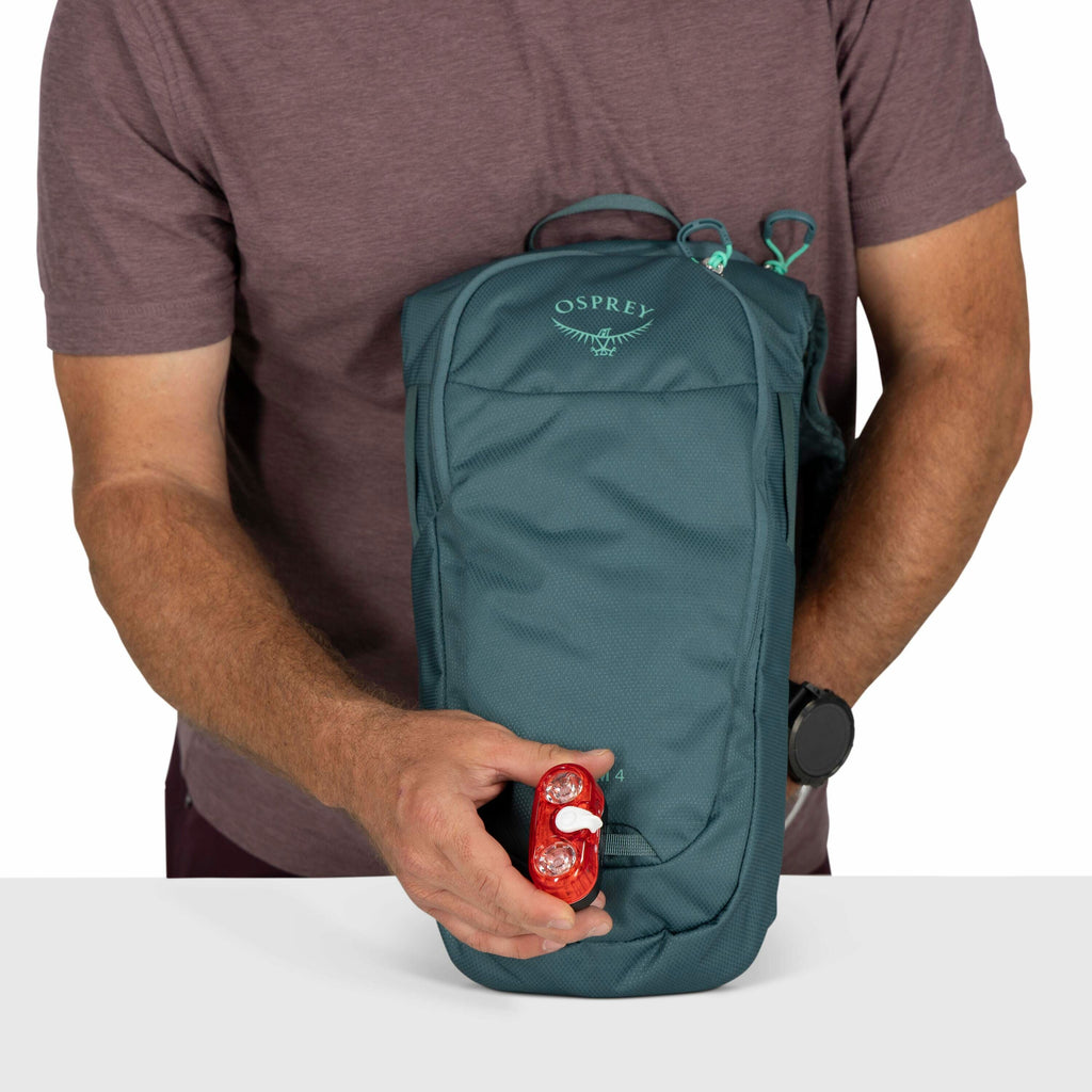 The image shows a person holding a small Osprey backpack in front of them. The backpack is a muted teal color and prominently displays the Osprey logo, featuring the brand name and an illustrated osprey bird, near the top. The individual is wearing a shor