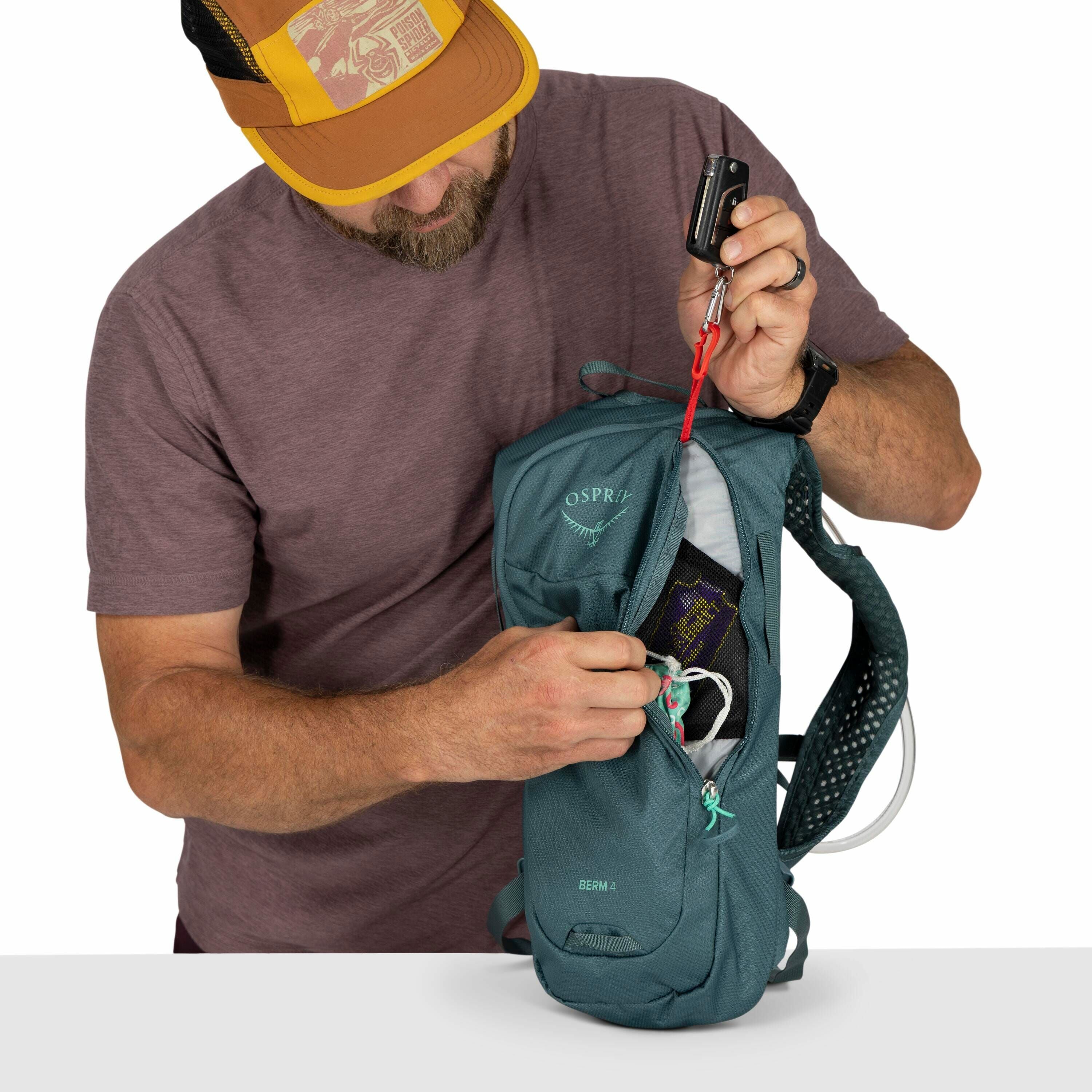 The image shows a person wearing a purple shirt and a yellow and black cap, holding a compact grey Osprey backpack. The backpack, labeled "BERM 4," is partially open, revealing a small mesh pocket containing what appears to be a snack bar or small item. T