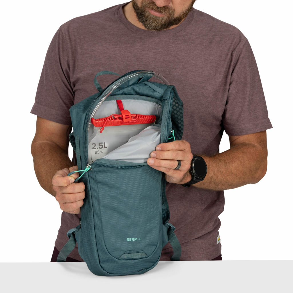 The image shows a man wearing a maroon t-shirt and demonstrating a teal-colored backpack with the brand name "BERM 4" visible at the bottom of the pack. He has the front compartment of the backpack open to reveal an internal hydration reservoir labeled "2