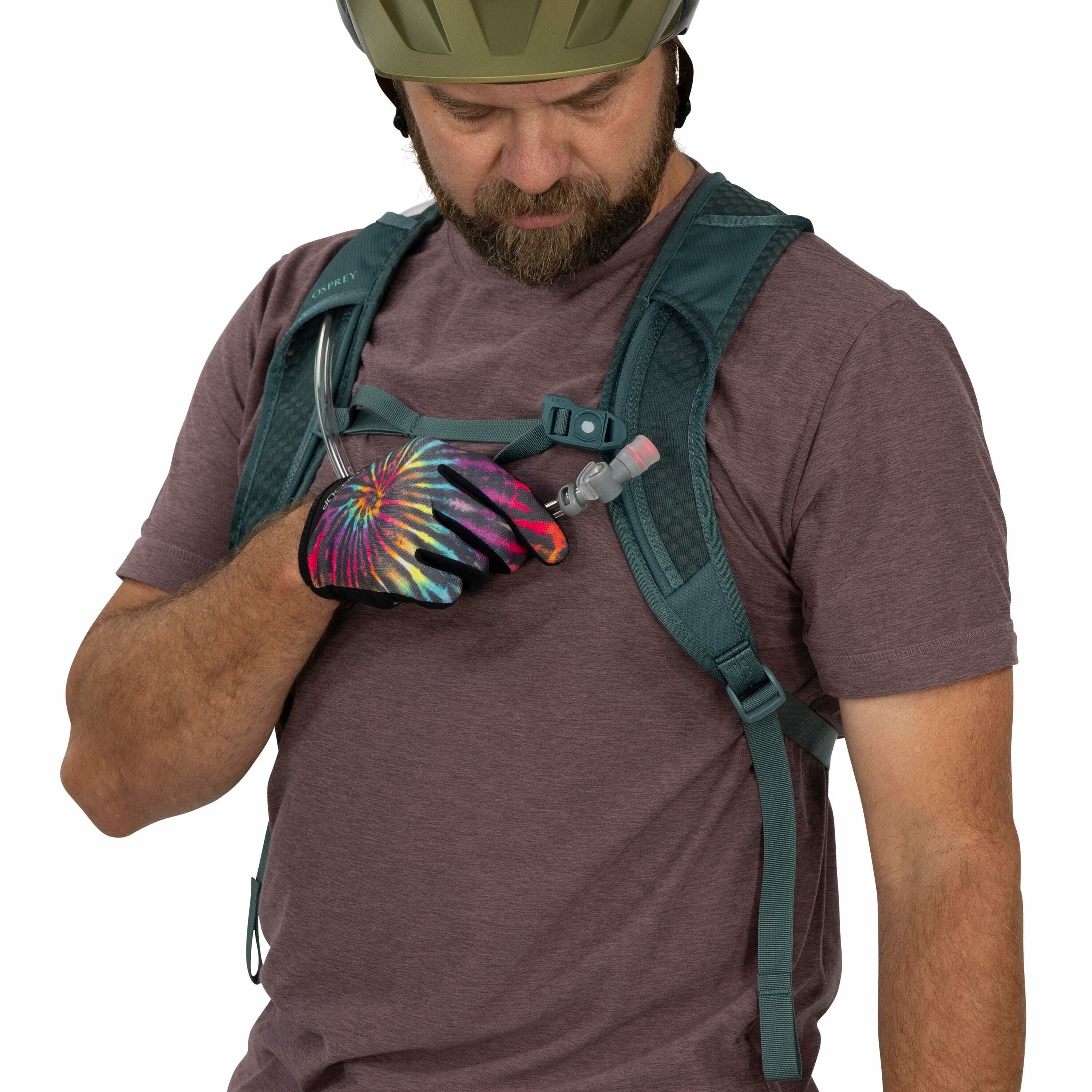 The image shows a person wearing a helmet, a maroon short-sleeve shirt, and an Osprey backpack with hydration features. The individual is holding the hydration tube with their right hand, which is clad in a vibrant tie-dye glove. The Osprey backpack is gr