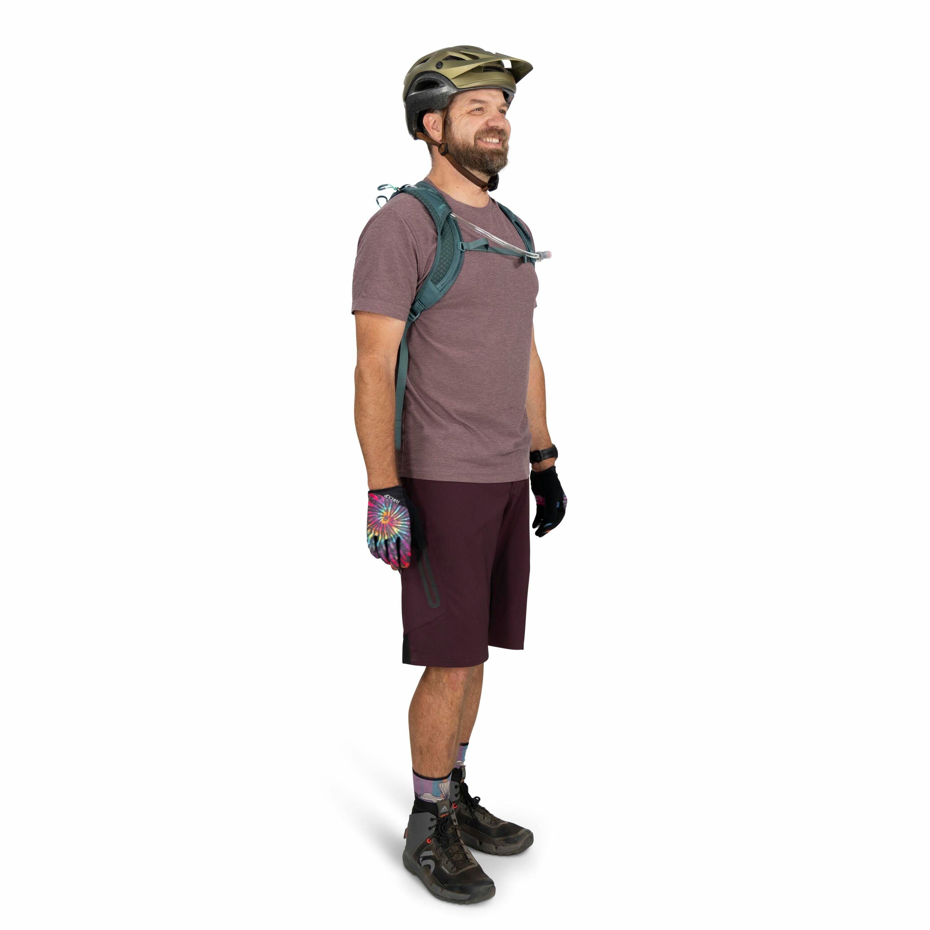 The image features a person dressed in biking attire, including a short-sleeve shirt and knee-length shorts in shades of purple. They are equipped with a green helmet for safety, and a backpack with a visible hydration tube, suggesting preparation for a l