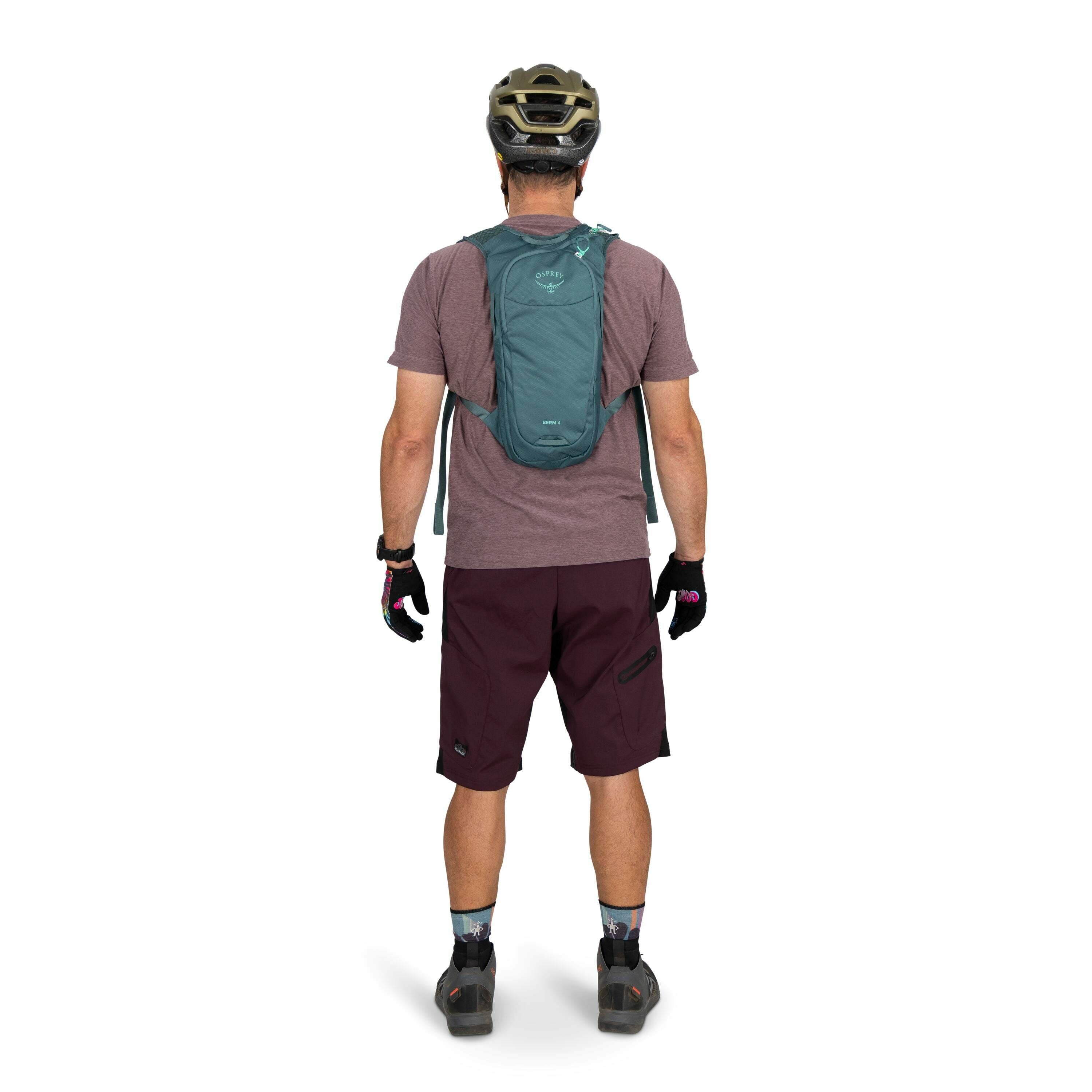 The image depicts a man standing with his back to the camera, dressed in cycling gear. He is wearing a helmet and has a small, lightweight Osprey backpack on his back. His outfit consists of a maroon t-shirt and dark maroon cycling shorts. He is also wear
