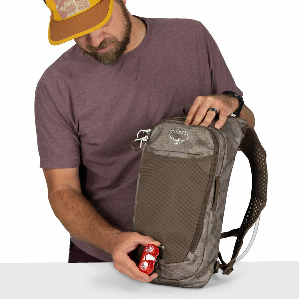 The image shows a person wearing a purple t-shirt and a yellow baseball cap while holding a grey and brown Osprey backpack. The backpack has a sleek, compact design with adjustable straps and multiple zippered compartments. The individual is holding a sma