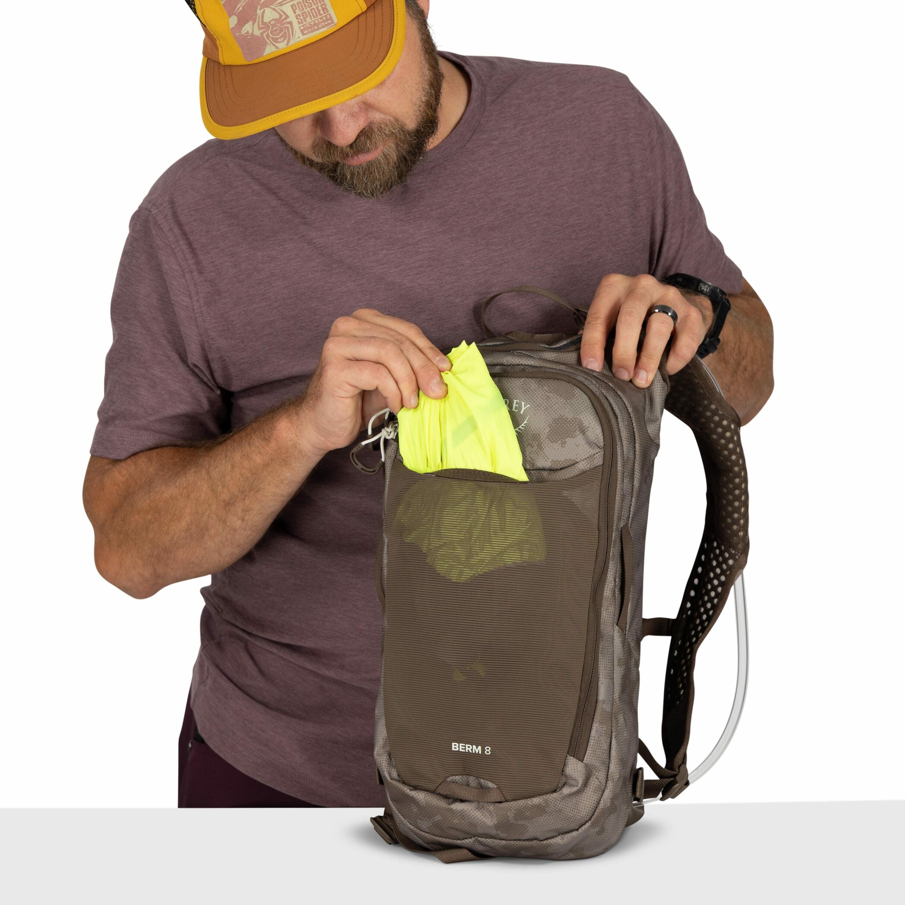 The image shows a person dressed in a purple t-shirt placing a bright yellow item into a gray backpack. The backpack, an Osprey brand, specifically the Berm 8 model, has a mesh front pocket where the yellow item is being stored. The person is wearing a bl
