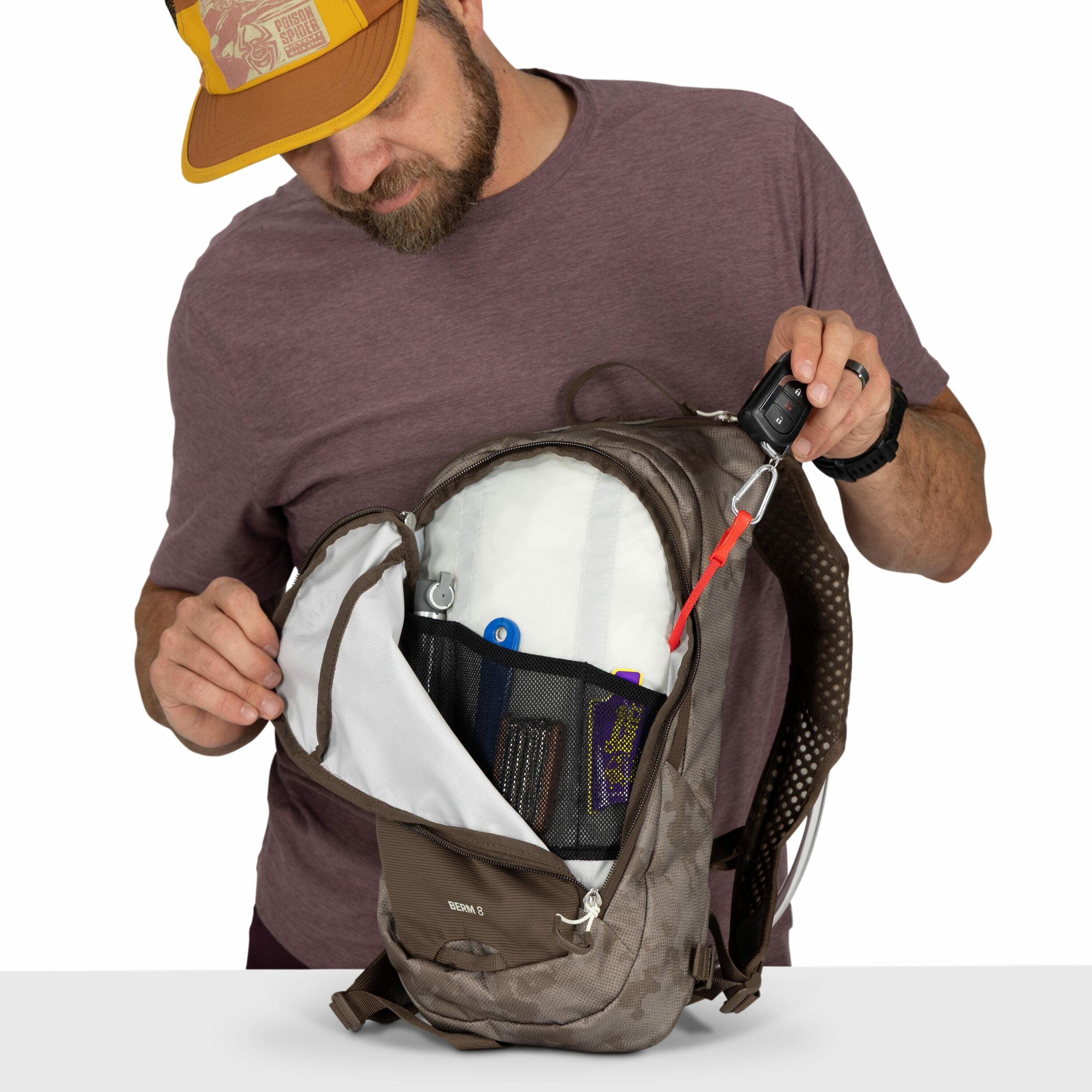 The image shows a person holding open a brown backpack, revealing its contents. The person is wearing a maroon t-shirt and a brown and yellow cap. The inside of the backpack is neatly organized with various items visible in mesh pockets, including a blue