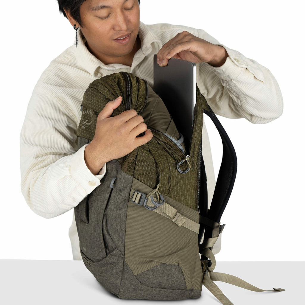 In the image, a person is placing a laptop inside a backpack. The backpack is two-toned with shades of olive green and gray and is made by Osprey, as indicated by the logo on its side. The person is wearing a long-sleeved, textured, white button-up shirt