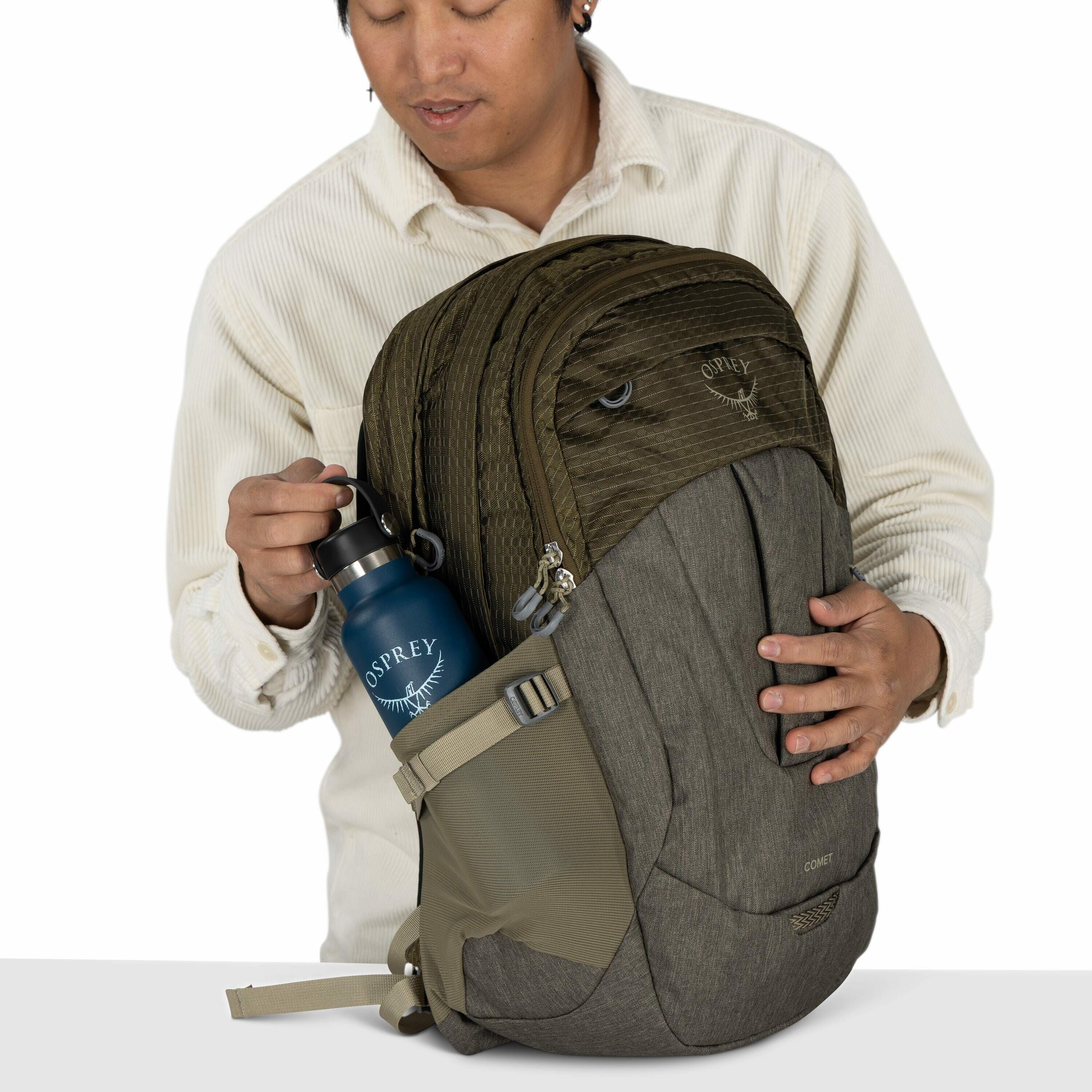 The image depicts a person holding an Osprey backpack, which is primarily green with a grey lower section. The backpack is quite large and features multiple zippered compartments. The person is wearing a cream-colored, textured long-sleeve shirt and is pl