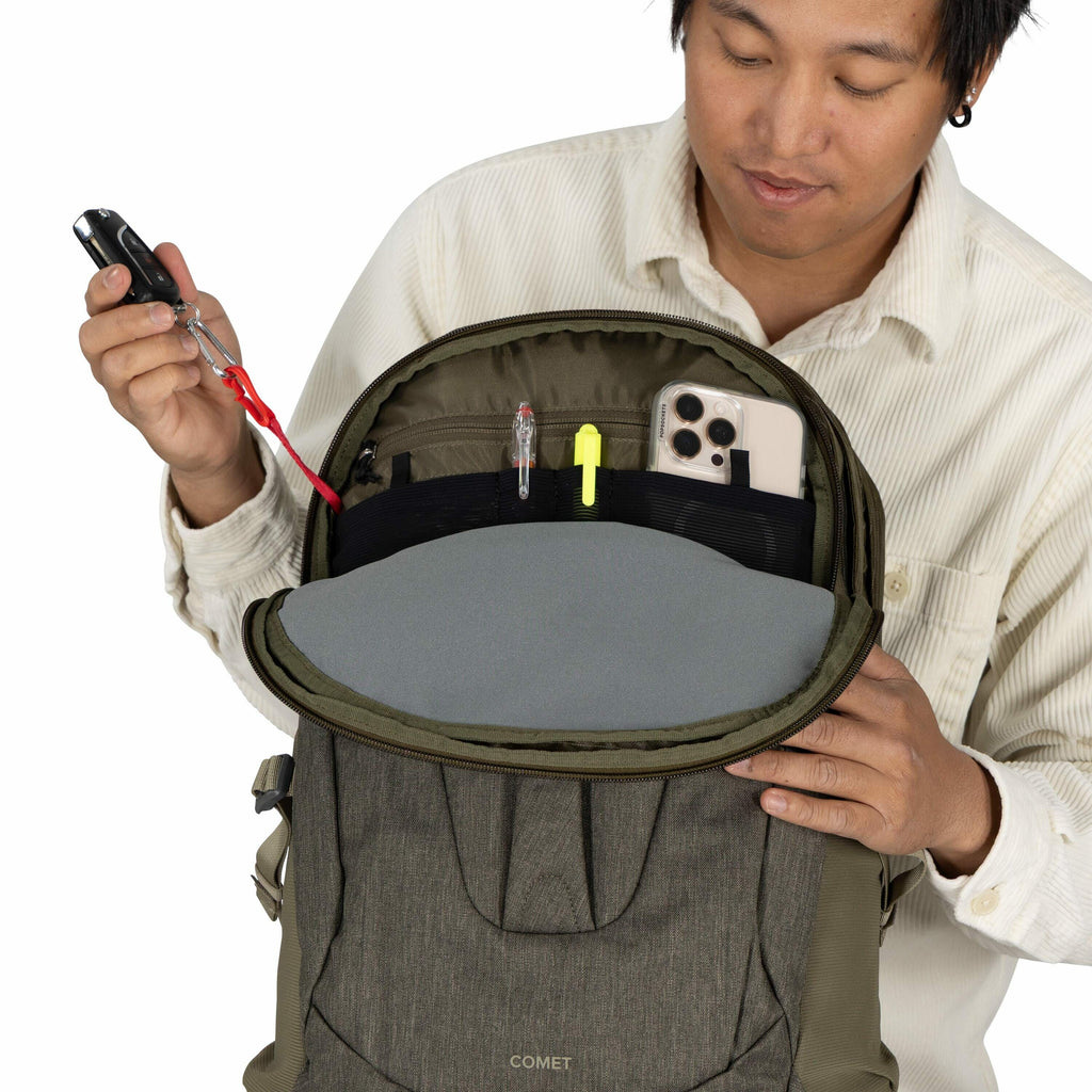 The image shows a person holding an open green and gray backpack with the brand name "COMET" visible on the front. The interior of the backpack is organized with various items: a smartphone in one pocket, a pen, and a highlighter in others. The individual