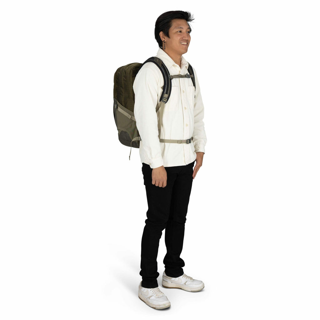 The image depicts a person standing and wearing a backpack. The individual is dressed in a white button-up shirt paired with black pants and white sneakers. The backpack appears to be dark green with a black strap and a chest buckle for support. The perso