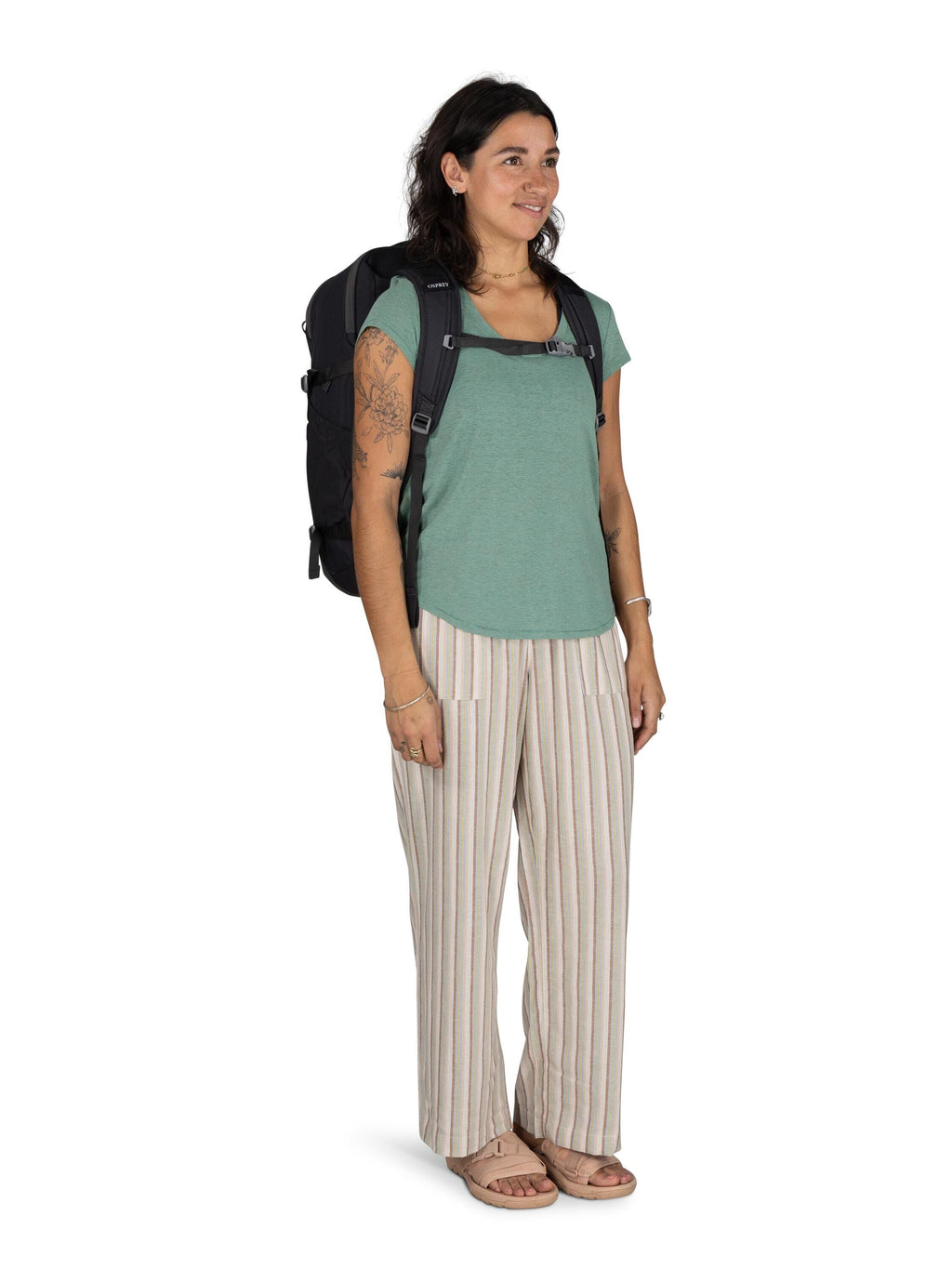The image shows a woman standing while wearing a casual outfit. She has a backpack strapped on her back, suggesting she might be traveling or commuting. Her attire consists of a teal-colored T-shirt paired with loose-fitting, striped pants. The pants have