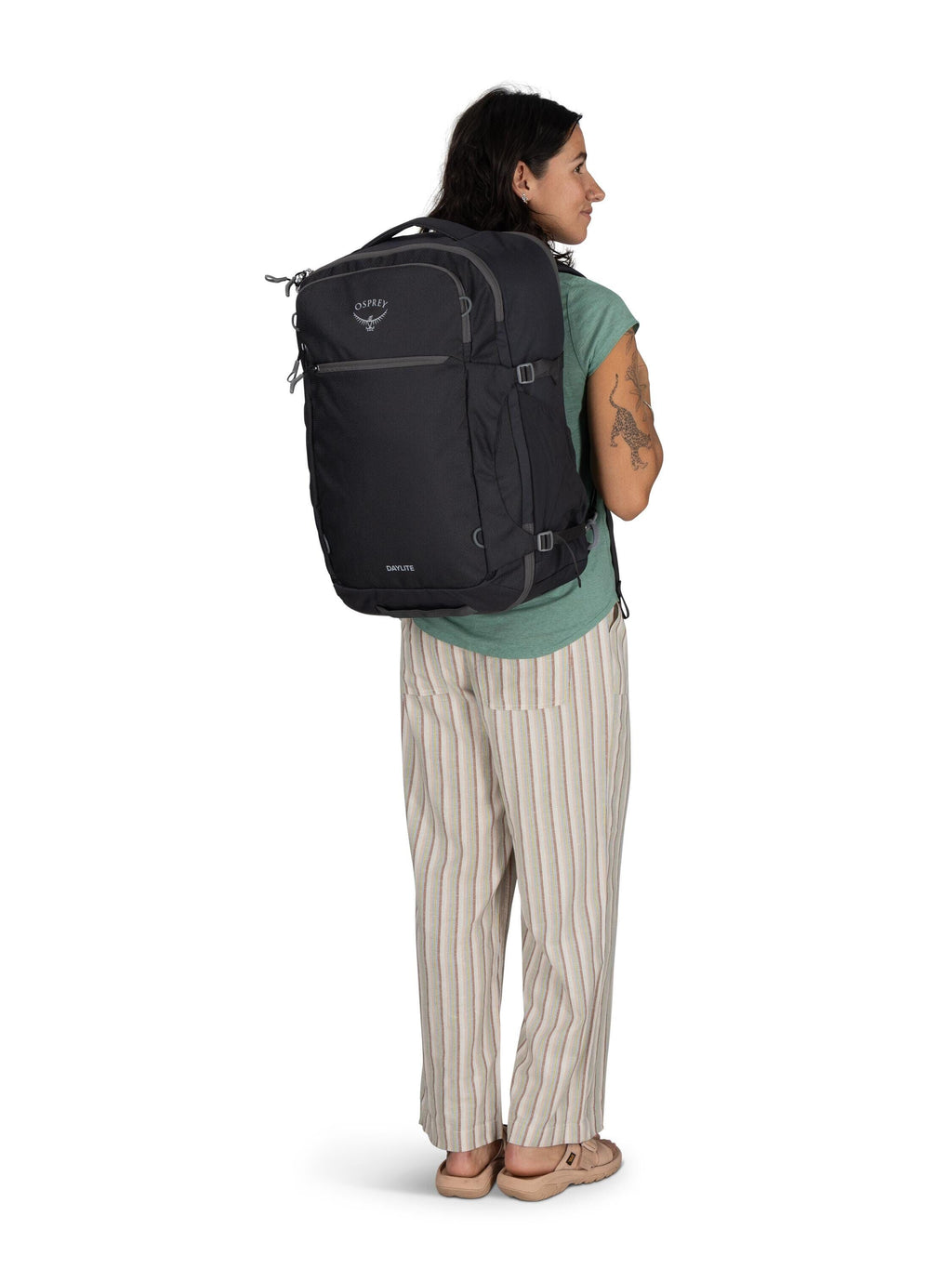 The image features a person standing and wearing a large black backpack. The backpack appears to be a travel or hiking backpack, designed for carrying substantial amounts of gear or belongings, and has several compartments and adjustable straps. The perso