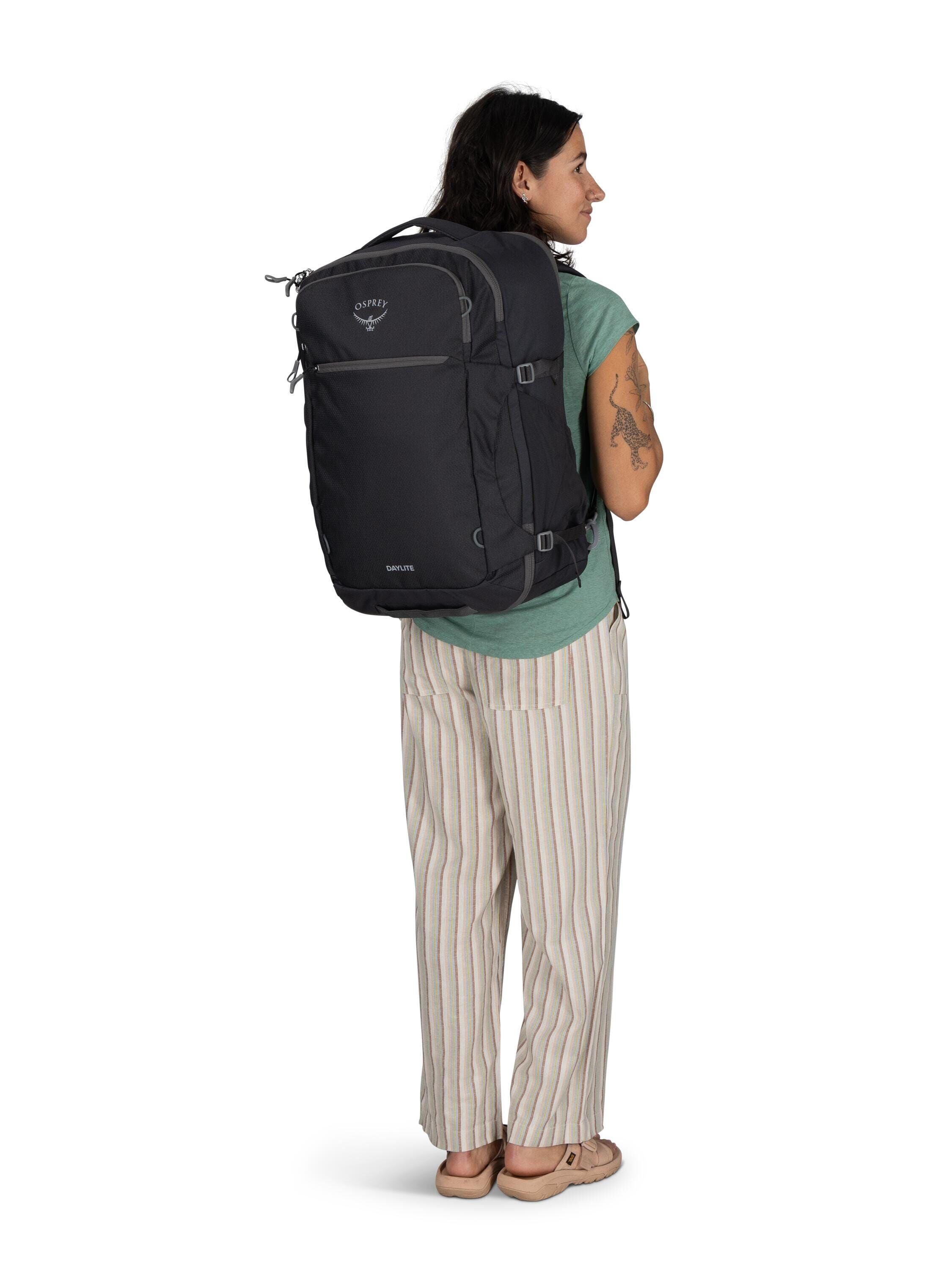 The image features a person standing and wearing a large black backpack. The backpack appears to be a travel or hiking backpack, designed for carrying substantial amounts of gear or belongings, and has several compartments and adjustable straps. The perso